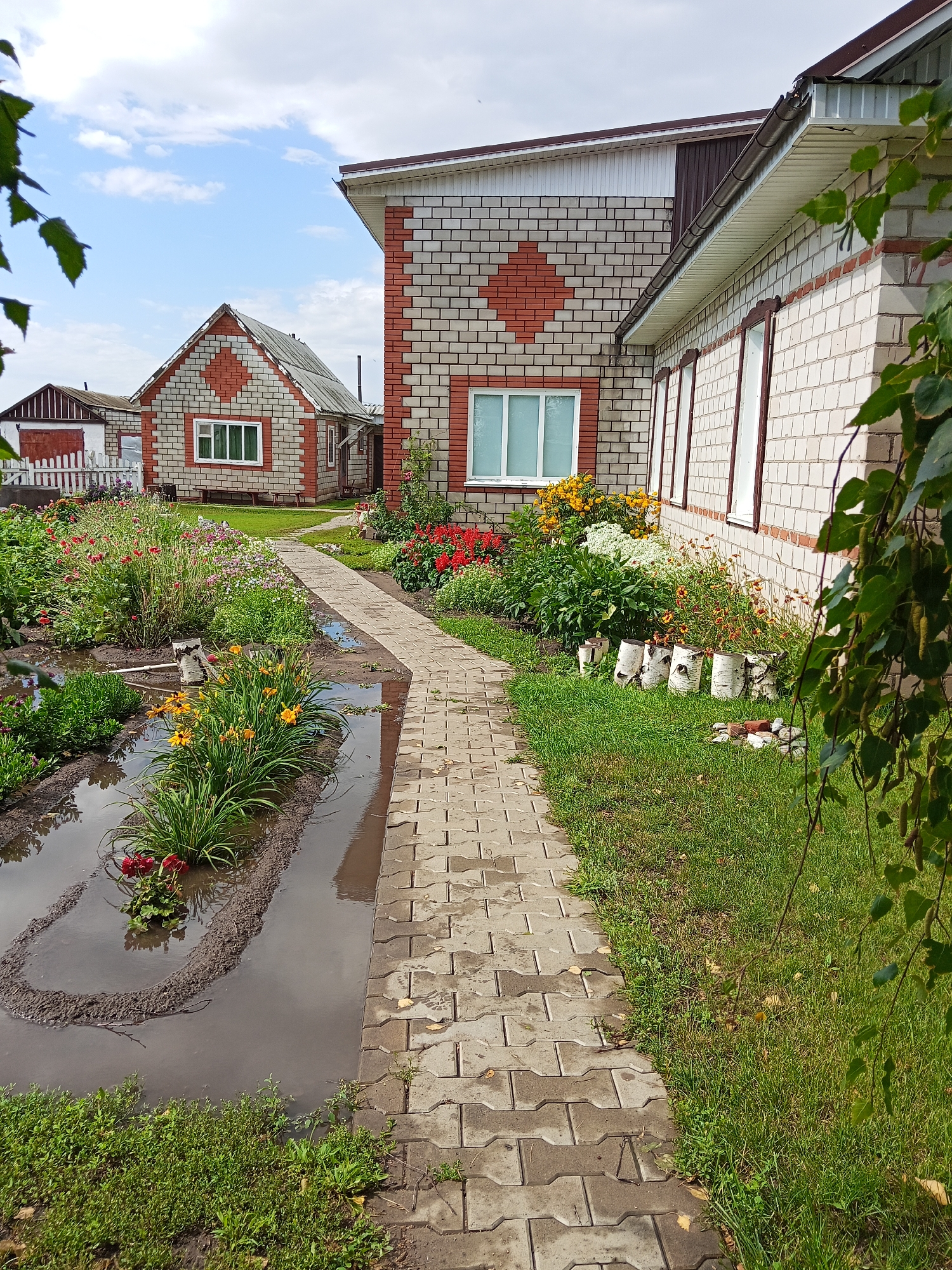 The image depicts a courtyard scene in the city of Khoroshee, Russia, dominated by two brick houses and a lush garden. A paved pathway, made of interlocking grey stones, runs diagonally through the frame, leading towards the houses. The pathway is partially submerged in water, suggesting recent rainfall.
To the left of the pathway is a vibrant garden filled with a variety of flowers, including red poppies and yellow daylilies. The garden bed itself is outlined with dirt and also contains standing water. A small wooden log and several birch logs are placed as decorative elements among the plants.
The larger house, to the right of the pathway, is constructed with light-colored bricks and features red brick accents arranged in a diamond pattern on its facade. It has a dark brown roof and several windows with white frames. The smaller house, further in the background and to the left, is also brick, with a similar diamond pattern in red and light bricks, and a grey roof. Another smaller building with a dark red door is visible behind the garden.
The environment is green and appears to be a residential area, possibly a dacha or country house setting, given the garden and the style of the houses. The sky is partly cloudy with patches of blue. The overall impression is one of a well-maintained, albeit waterlogged, outdoor space. There are no people visible in the image.