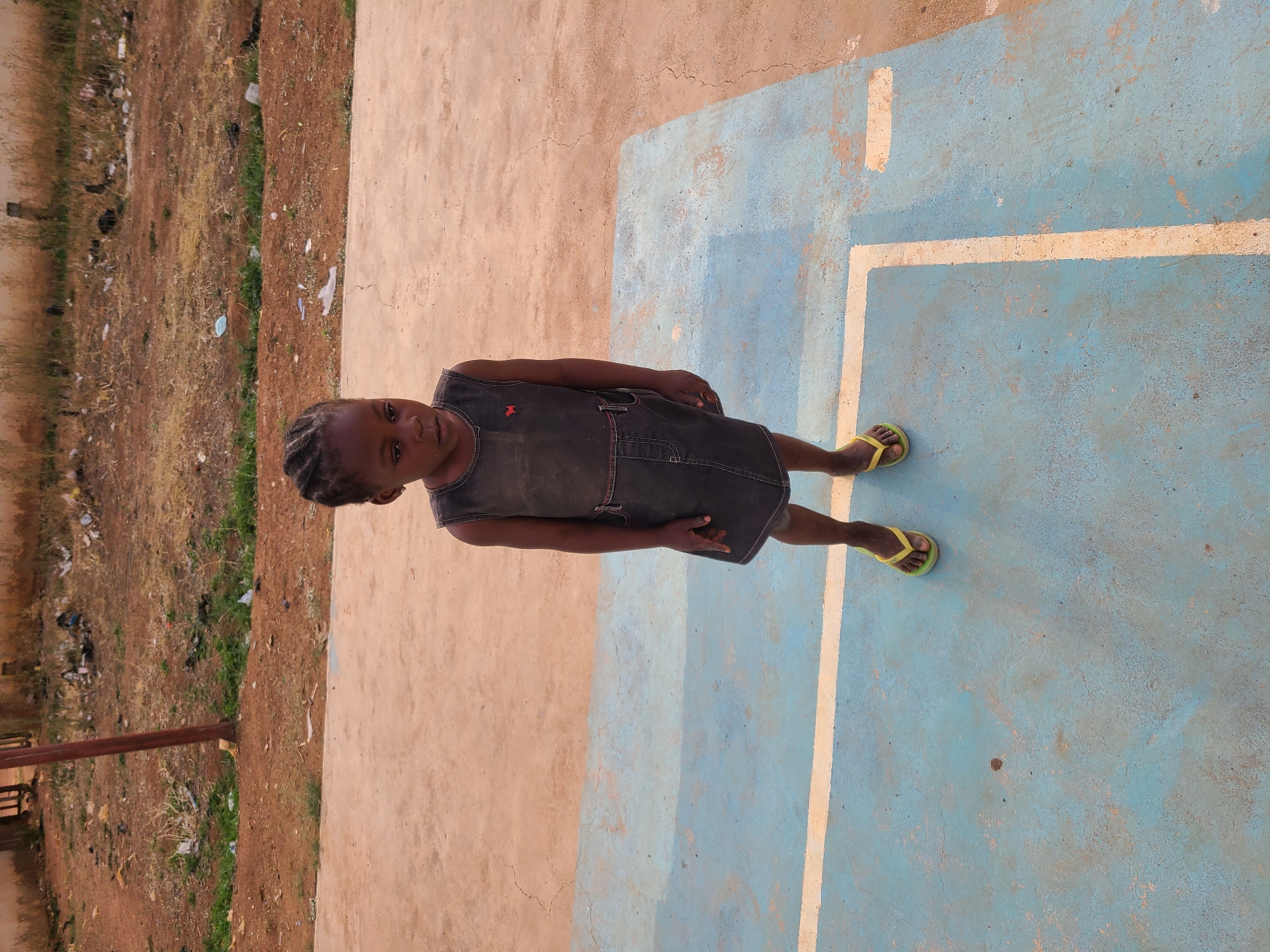 A young girl stands in an outdoor setting in Jos, Nigeria. She has dark skin and her hair is styled in braids. She is wearing a dark, sleeveless dress, possibly made of denim, which features reddish-brown stitching along the seams and a small red star emblem on the chest area. Her feet are clad in bright yellow flip-flops.
The girl is positioned on a paved surface that is partially painted blue with worn white lines, suggesting a sports court or recreational area. This blue area transitions into a lighter beige-colored concrete or paved surface behind her to the left. Beyond the paved area, a strip of reddish-brown dirt is visible, covered with dry grass and some sparse green vegetation. Scattered litter, including plastic bags and other debris, is present on the ground in this unkempt background. A dark, possibly wooden or metal, pole stands upright in the dirt area to the far left.
The lighting indicates bright daylight, with clear conditions. The girl looks directly at the camera with a neutral expression. No specific activity is taking place other than her standing for the photograph. There is no visible text in the image.