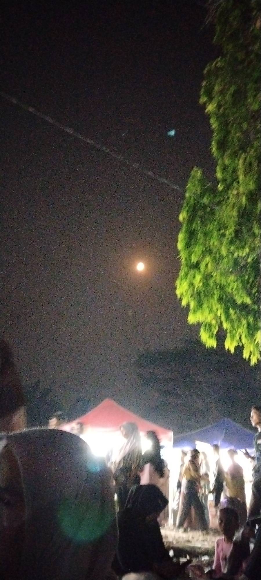 An outdoor night scene in Sido Rahayu, Indonesia, captures a gathering under a hazy, moonlit sky. The moon, a pale orange orb, is visible in the upper center of the frame, surrounded by a faint glow. To the right, the dense, green foliage of a large tree dominates the upper right quadrant. In the middle ground, several tents are illuminated from within, casting a warm light onto the people gathered around them. The foreground is blurred, with indistinct figures and objects suggesting a busy, crowded environment. People are seen in various states of activity, some standing, some seated, and the overall impression is of a community event or market. The atmosphere appears lively and somewhat diffused due to the low light conditions.