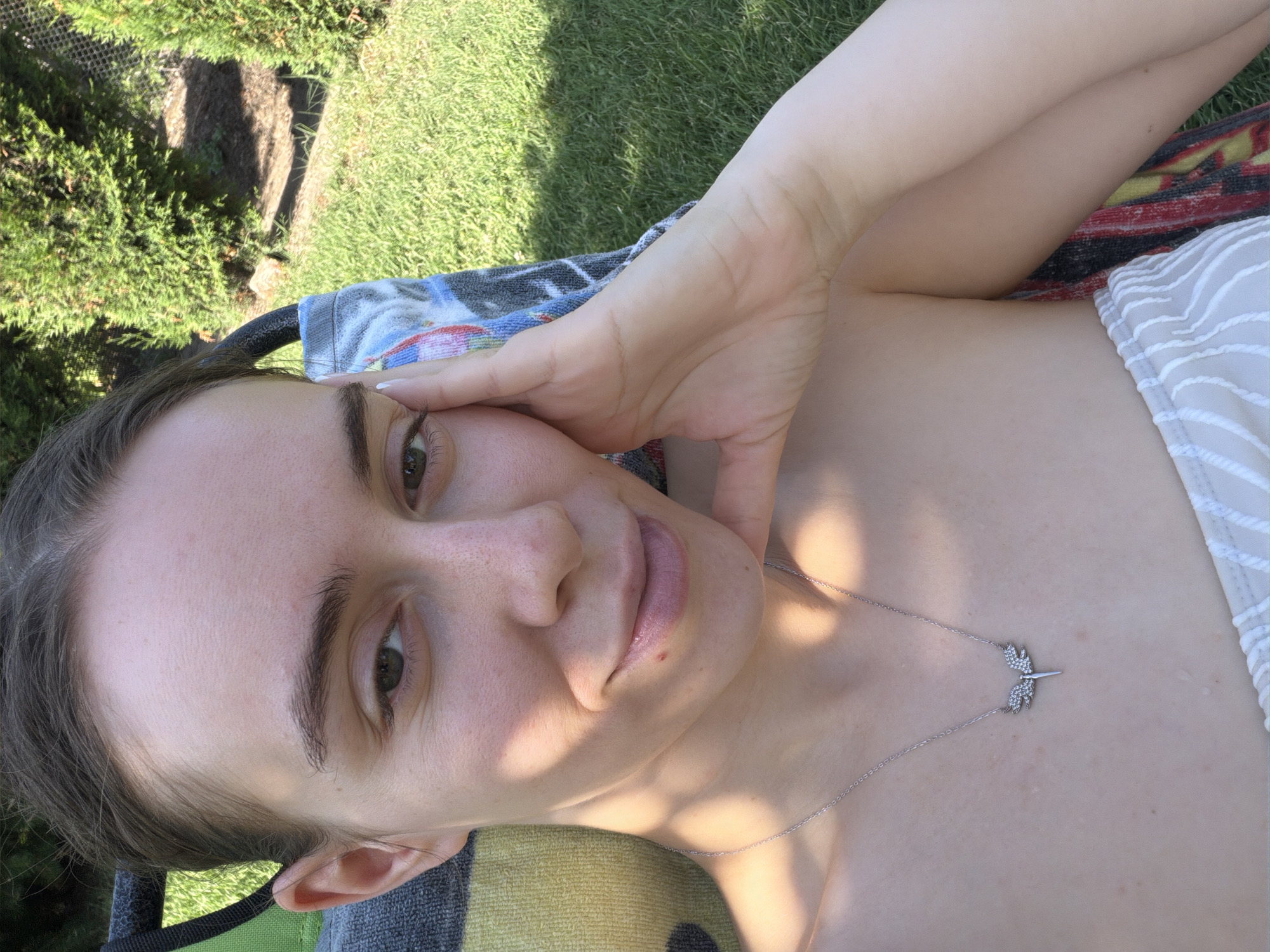 A young woman with light skin and dark hair is resting outdoors on a sunny day. She is lying down, propped up by a colorful striped blanket and a chair. Her left hand is resting on her cheek, and she is looking at the camera. She is wearing a silver necklace with a dragonfly pendant. The background consists of green grass and trees, suggesting a park or backyard setting. The lighting indicates it is daytime. The woman appears relaxed and is positioned within the city limits of Bratanitsa, Bulgaria.