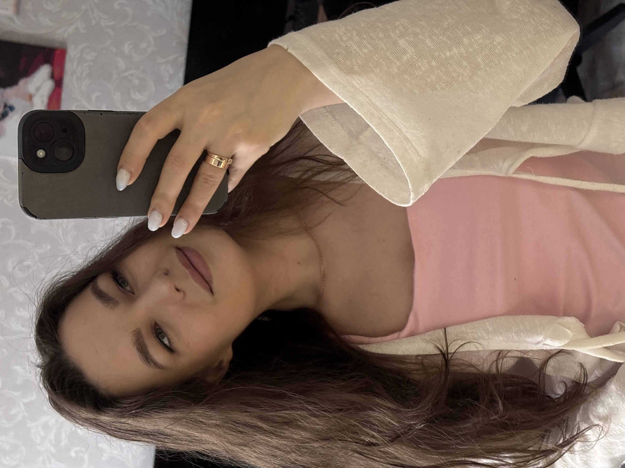 A young woman is taking a selfie with a smartphone. She is wearing a pink top and a cream-colored linen-like cardigan. Her hair is long and brown. She is wearing a gold ring on her left ring finger. The background is blurry but appears to be a textured wall. There is no indication of the city of Chaikovsky, Russia in the image.