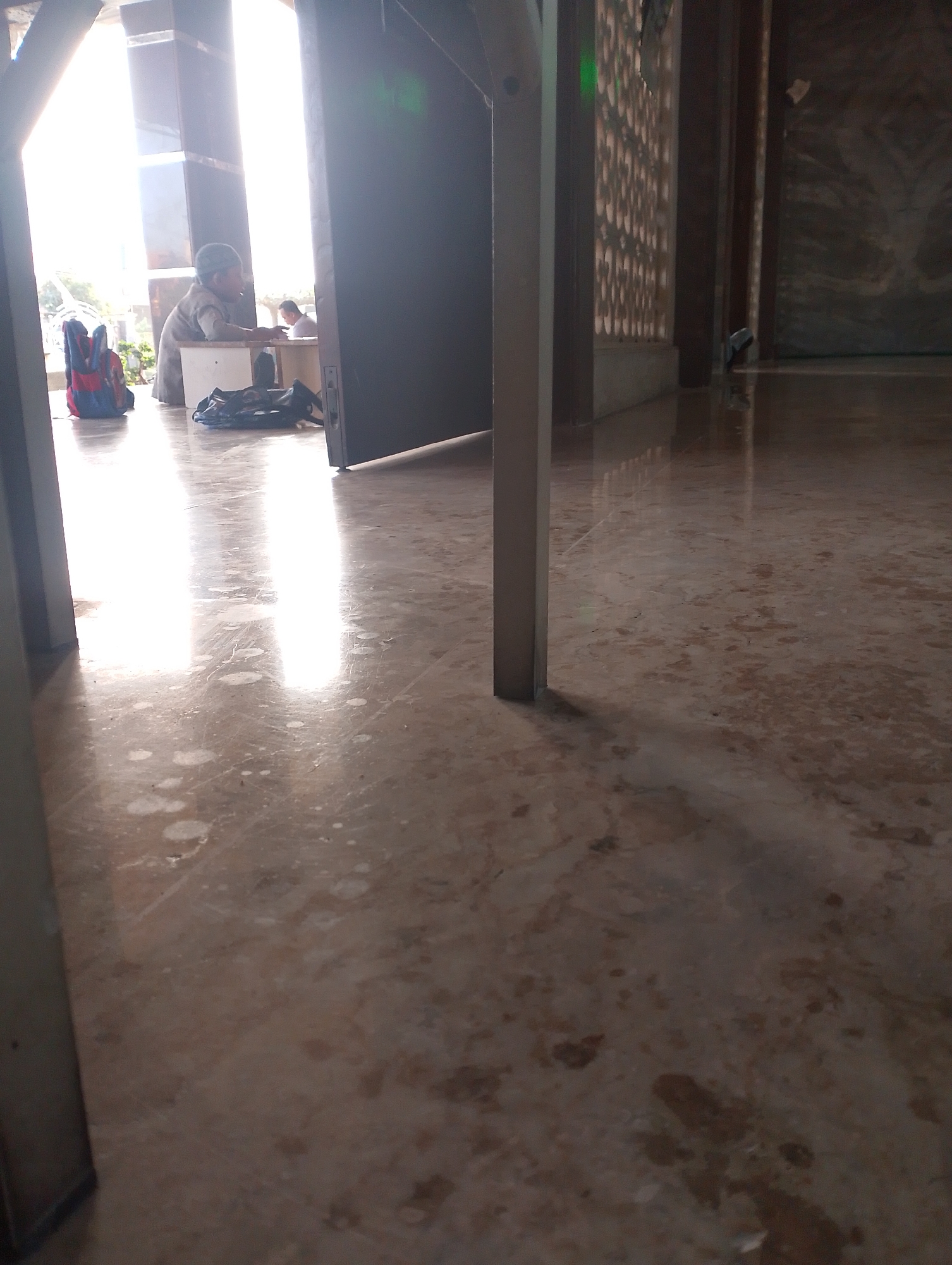 In Sukodono, Indonesia, a low-angle shot captures a brightly lit interior space. The floor, made of polished marble with brown and beige veining, reflects the light. In the background, a doorway opens to a bright exterior, with strong vertical light shafts illuminating the scene. To the left of the doorway, a child wearing a gray garment and a cap sits at a light-colored table. In front of the child, a backpack with red, blue, and black colors rests on the floor. A black bag is also on the floor, placed behind the table. To the right of the doorway, a patterned wall with square openings is visible. The overall atmosphere is quiet and serene.