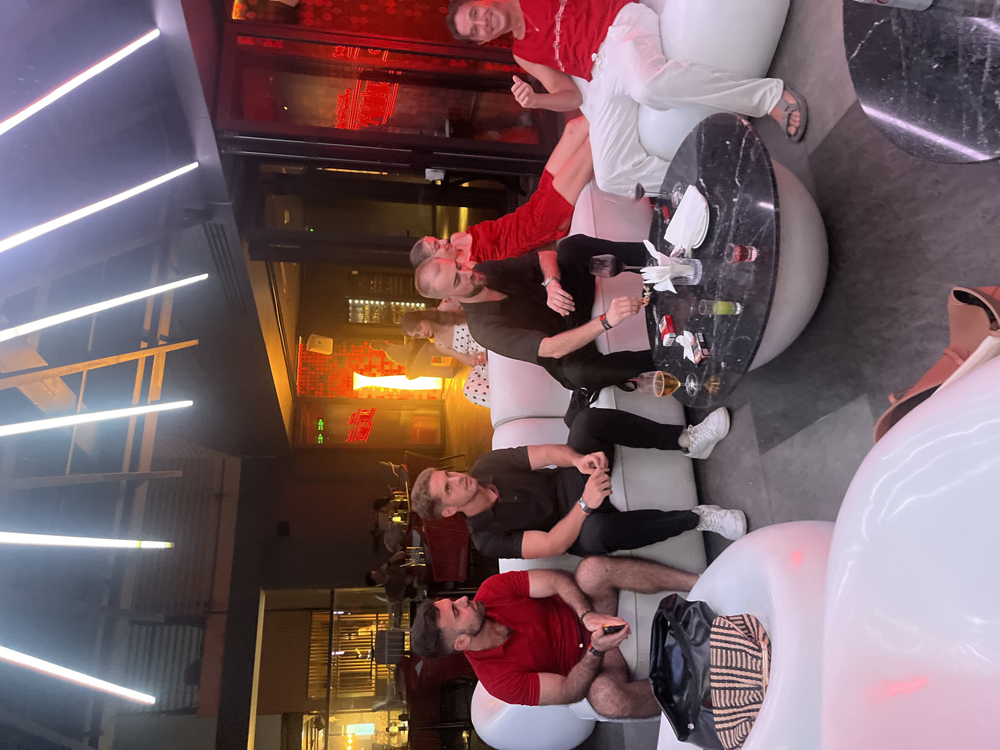 In an indoor setting that appears to be a lounge or bar, several people are seen relaxing and socializing. The scene is dimly lit, with prominent neon lights adding to the ambiance.
The subjects include a group of men and women seated on modern, white, bulbous couches. One man, wearing a red t-shirt, is seated on the right side of the frame, holding a phone. Another man, in a black t-shirt, is seated to his left, looking towards the side. Further up and to the left, another man in a black t-shirt is seated with a woman in a polka dot dress. At the top of the frame, a man in a red shirt is giving a thumbs-up, while a person in white pants is also visible.
On a black marble-top table in the center of the image, several glasses of drinks, cigarettes, and napkins are present, suggesting a casual gathering. The overall atmosphere seems relaxed and convivial. The lighting, particularly the neon signs, hints that it might be evening or nighttime.
The location is identified as Đà Nẵng, Vietnam. While specific weather is not discernible, the indoor setting suggests it's independent of external conditions. There is visible text on neon signs in the background, which appear to be in an Asian script, possibly Vietnamese given the location context. The red neon sign in the background has stylized lettering.