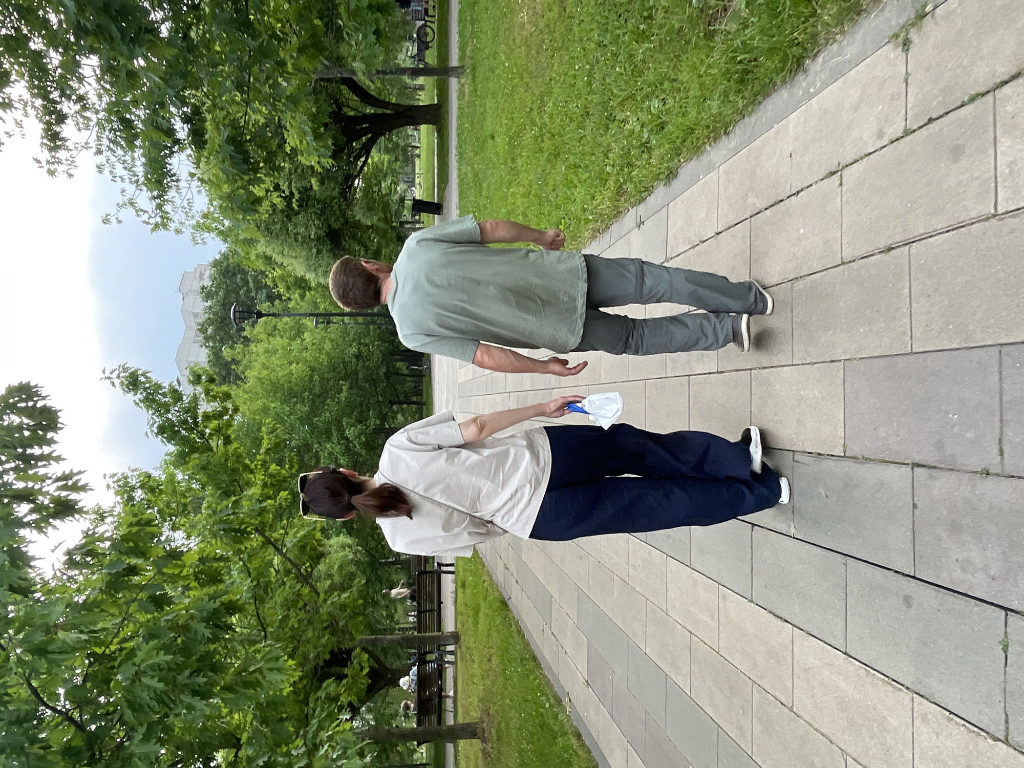 In Moscow, Russia, two individuals are walking away from the camera on a paved park path. The person on the left, a woman with dark hair pulled into a ponytail, wears a light-colored t-shirt and dark, wide-legged pants. She carries a white object, possibly a face mask, in her right hand. The person on the right, a man with short brown hair, is dressed in a light green t-shirt and grey pants. They are walking on a path made of rectangular stone slabs, bordered by a grassy area on the right. Lush green trees line both sides of the path, with more trees and some buildings visible in the background. The sky is overcast. In the distance on the left, several people are seated on park benches.