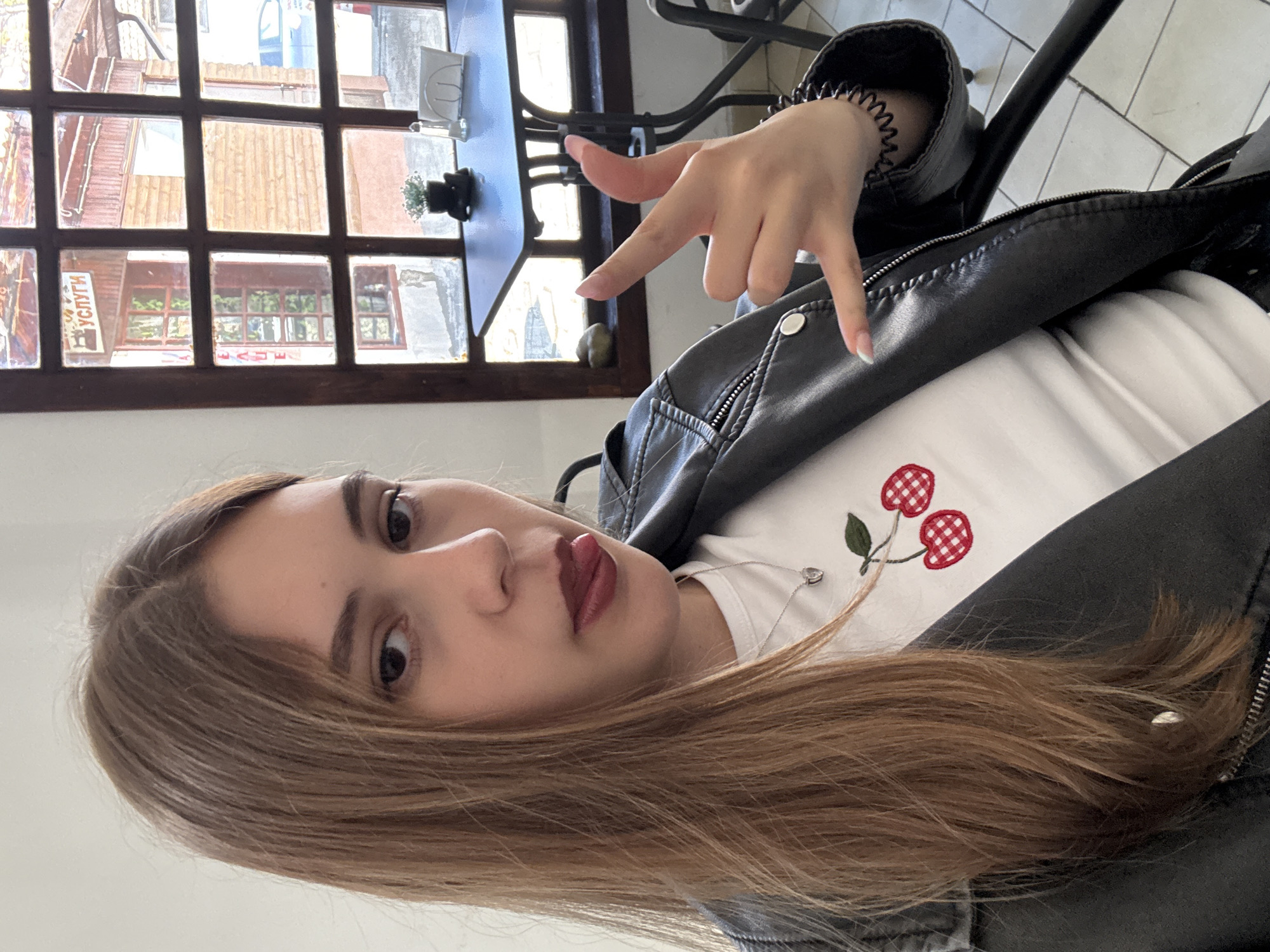 A young woman is depicted in the image. She has fair skin, brown hair, and is wearing a black leather jacket over a white shirt with a cherry design. She is holding her hand up making a "rock-on" sign. The location seems to be a cafe or restaurant, based on the table and window in the background. Through the window, the cityscape of Panagyurishte, Bulgaria is visible. The structures have a tiled roof and the window seems to have a sign hanging at the top left.