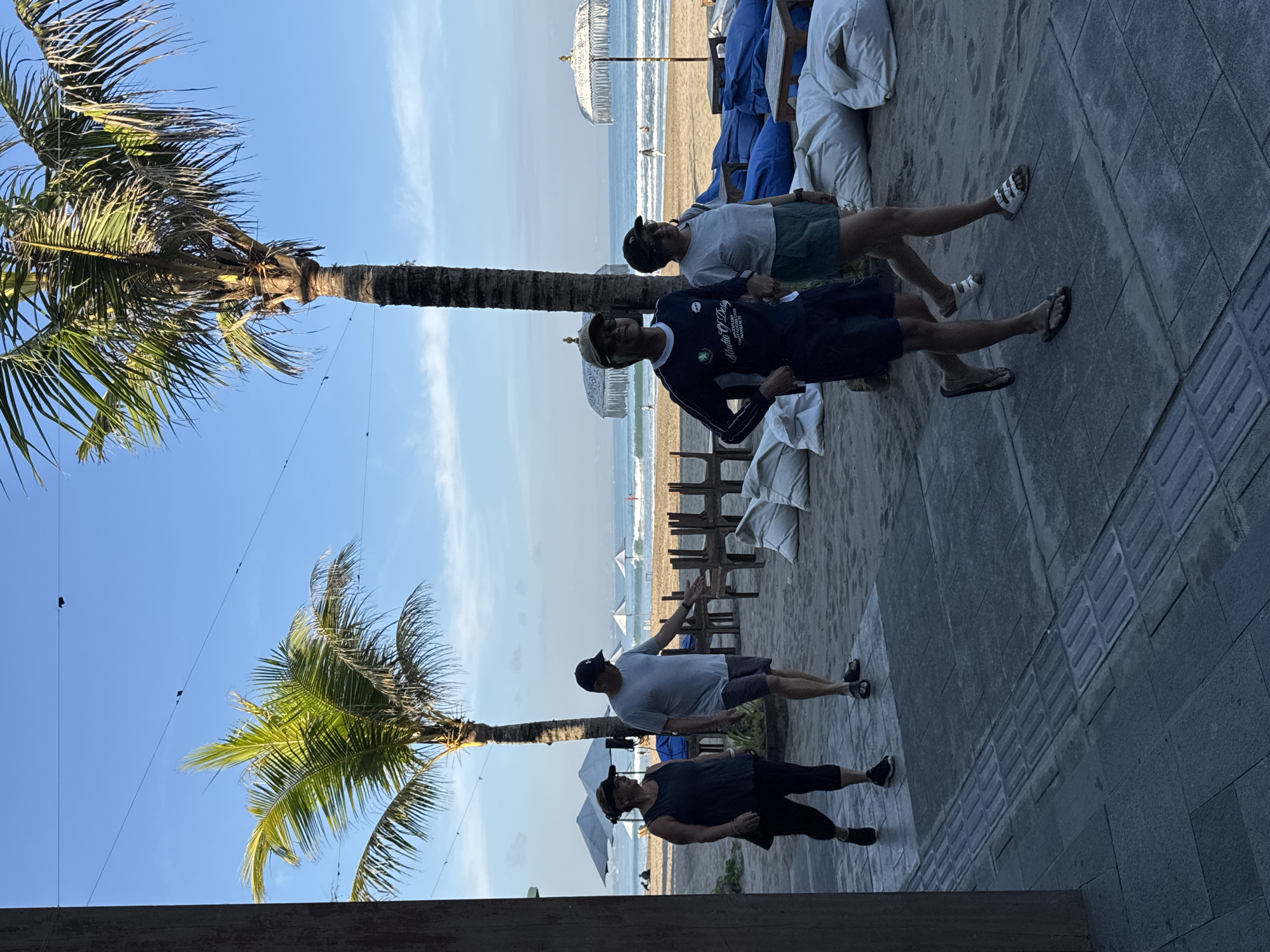 This image captures a vibrant, sunny beach scene in Kuta, Indonesia, featuring a paved walkway adjacent to the sand and ocean. Two prominent palm trees with dense green fronds flank the left side, their trunks extending upwards against a clear blue sky.
On the paved walkway, four individuals are present, appearing relaxed and casually dressed. From left to right (as the image is viewed upright): one person is dressed in a dark top, dark capri pants, and black shoes, possibly wearing a visor; next, a man in a light grey t-shirt, dark shorts, a black baseball cap, and dark flip-flops stands with one arm slightly raised. To his right, another man wears a long-sleeved dark blue shirt with the text "STUDIO G DESIGN" and "INTERNATIONAL" visible on it, a light-colored cap, dark shorts, and dark flip-flops. The fourth person on the walkway, furthest to the right, wears a light grey t-shirt, dark shorts, a dark cap, and white Adidas sandals.
Beyond the walkway, the sandy beach extends towards the ocean, where gentle waves can be seen. Several white beach umbrellas and covered lounge chairs are scattered across the sand. In the distance, near the water's edge, a few indistinct figures are visible. The sky is bright blue with only a few wispy clouds, indicating clear, pleasant weather, likely late morning or early afternoon given the bright, direct sunlight. The scene conveys a leisurely and tranquil atmosphere typical of a tropical vacation spot.