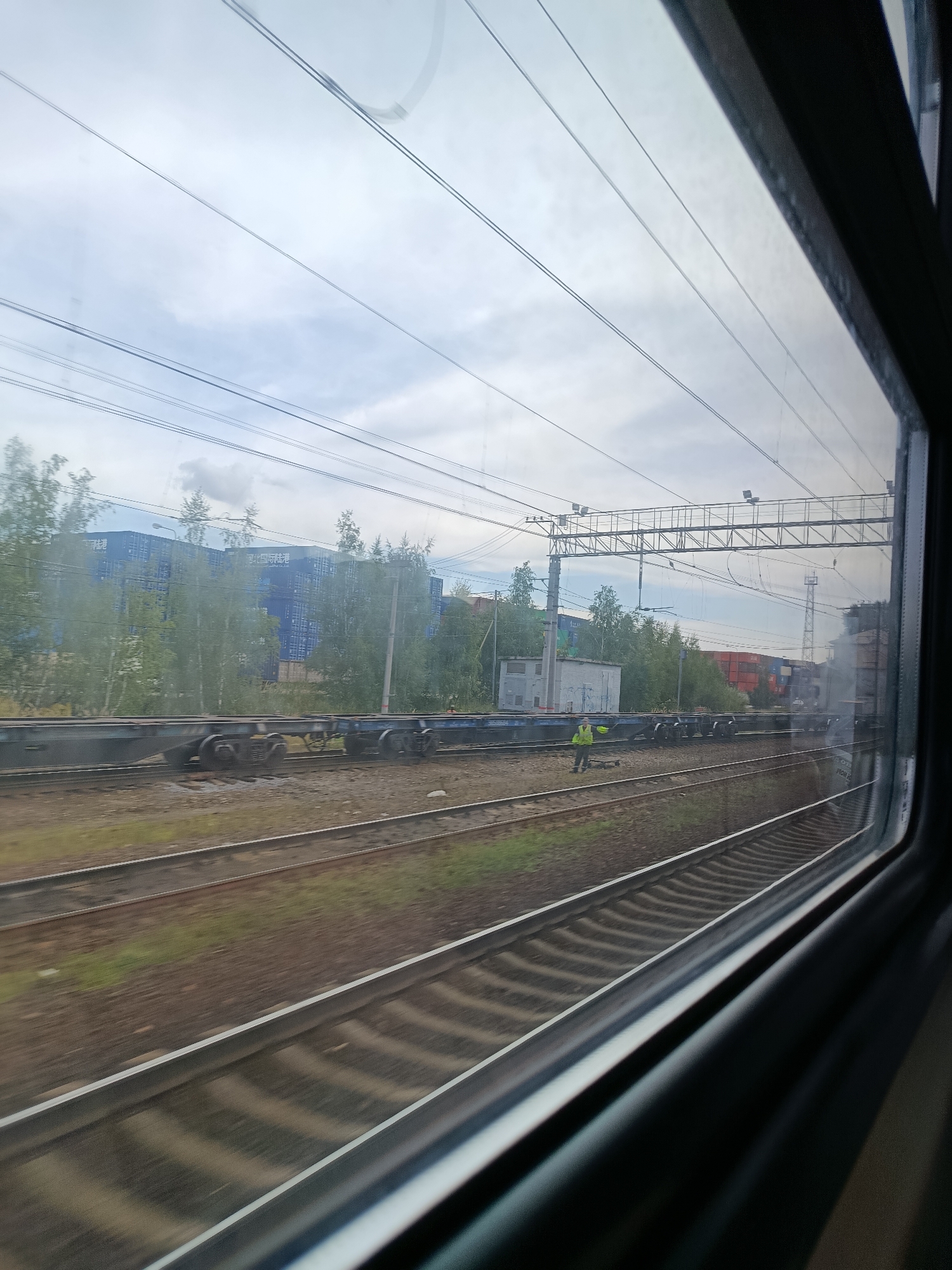 The image, taken from a train window, shows a railway track winding through an industrial area. In the background, a stack of blue shipping containers is visible behind a line of trees. A person wearing a high-visibility yellow vest stands on the tracks, appearing to be directing something or someone. Overhead power lines stretch across the sky, typical of a rail network. The scene is consistent with a view of the Elektrougli railway station in Elektrougli, Russia, an area known for its industrial and transportation infrastructure.