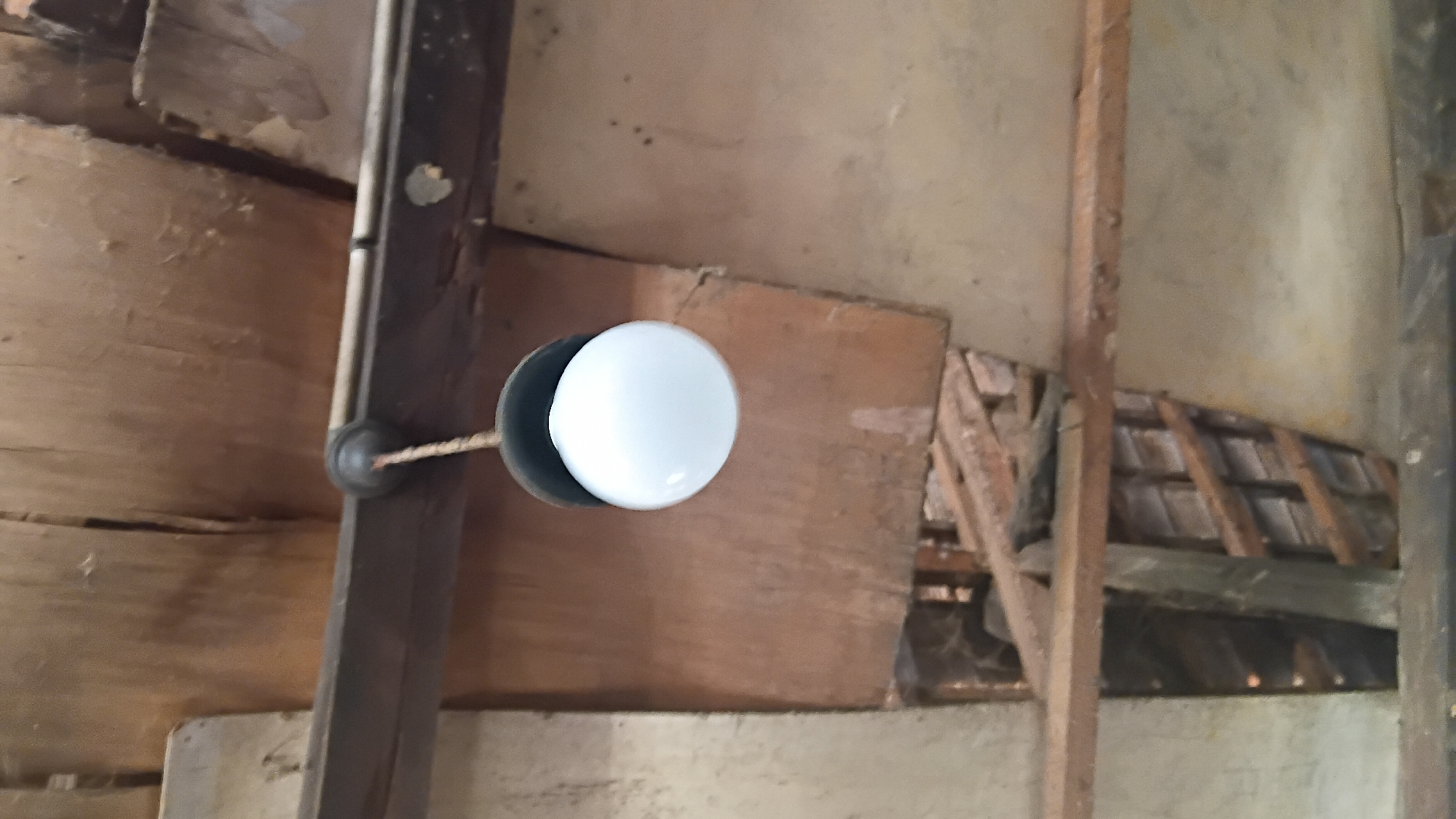 A single, white, spherical lightbulb hangs from a dark fixture by a braided cord, suspended from a wooden ceiling structure. The ceiling appears to be made of rough, unfinished wood panels and beams. The background reveals more of the same rustic, possibly dilapidated ceiling or roof construction, with visible gaps and structural elements. The image does not contain any people, animals, or identifiable landmarks. Without additional context, the specific location in Prabumulih, Indonesia cannot be determined from the visual information provided.