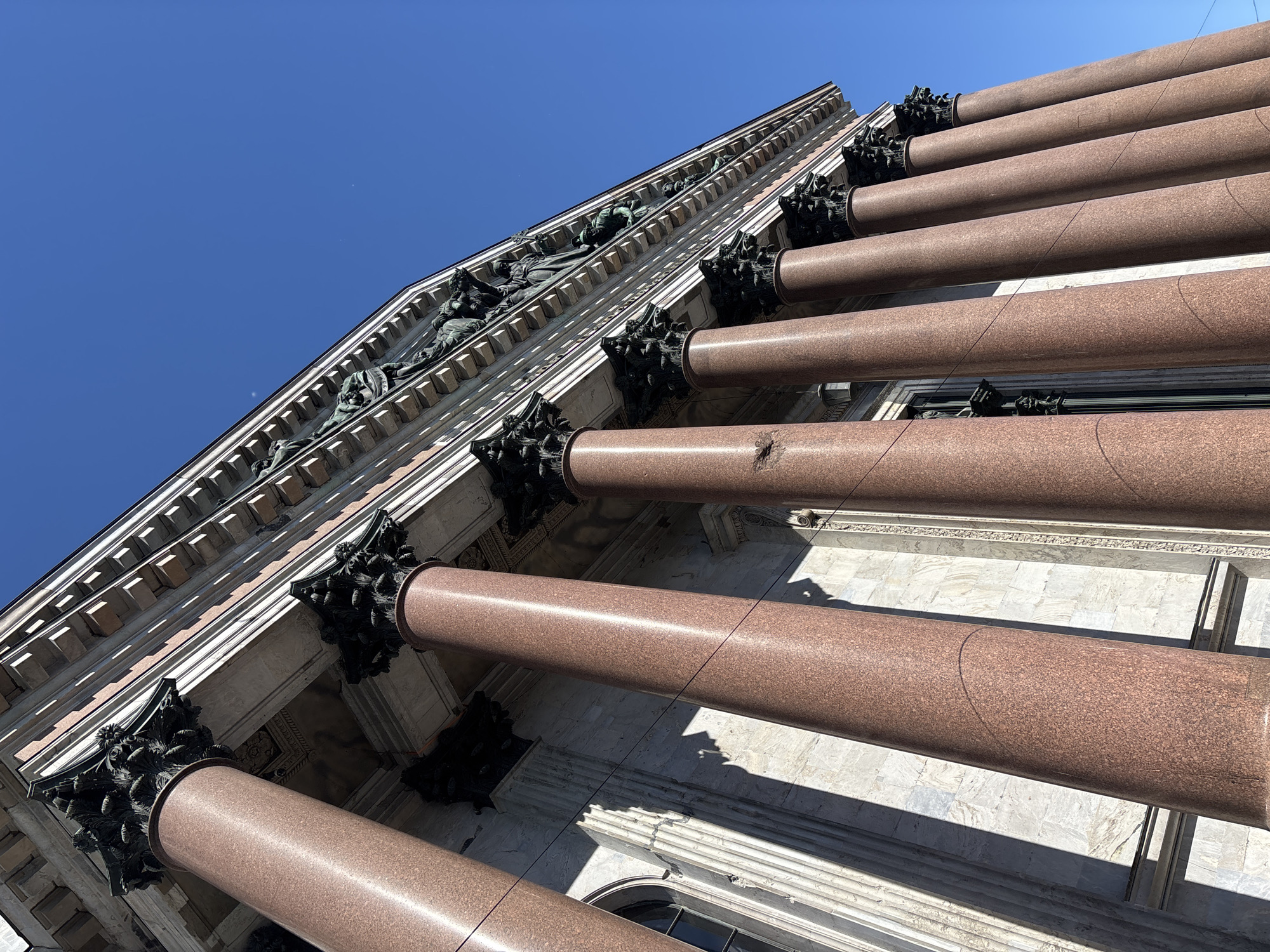 The image shows a low-angle view of the facade of Saint Isaac's Cathedral in Saint Petersburg, Russia. The facade is characterized by a series of tall, red granite columns with ornate black capitals. Above the columns, a frieze with sculpted figures is visible against a clear blue sky. The architectural style is classical. The lighting suggests a bright, sunny day, casting shadows from the columns onto the building's facade. There are no people visible in the image.
