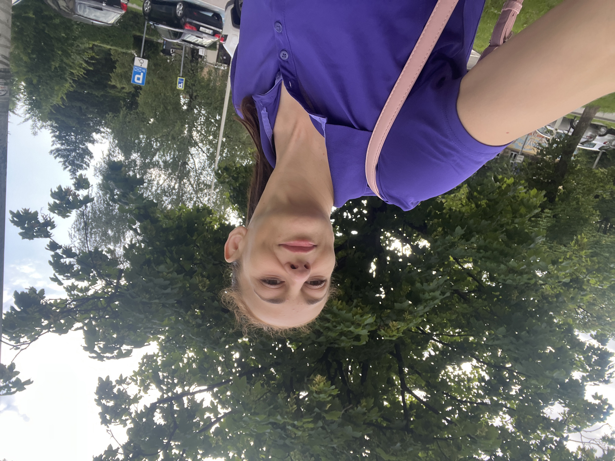 A woman takes a selfie outdoors, with lush green trees behind her and a cloudy sky above. She has her hair pulled back and is wearing a purple collared shirt and a pink strap across her body. In the background, cars are parked on the side of a street. Two blue parking signs, one with a wheelchair symbol, are visible.