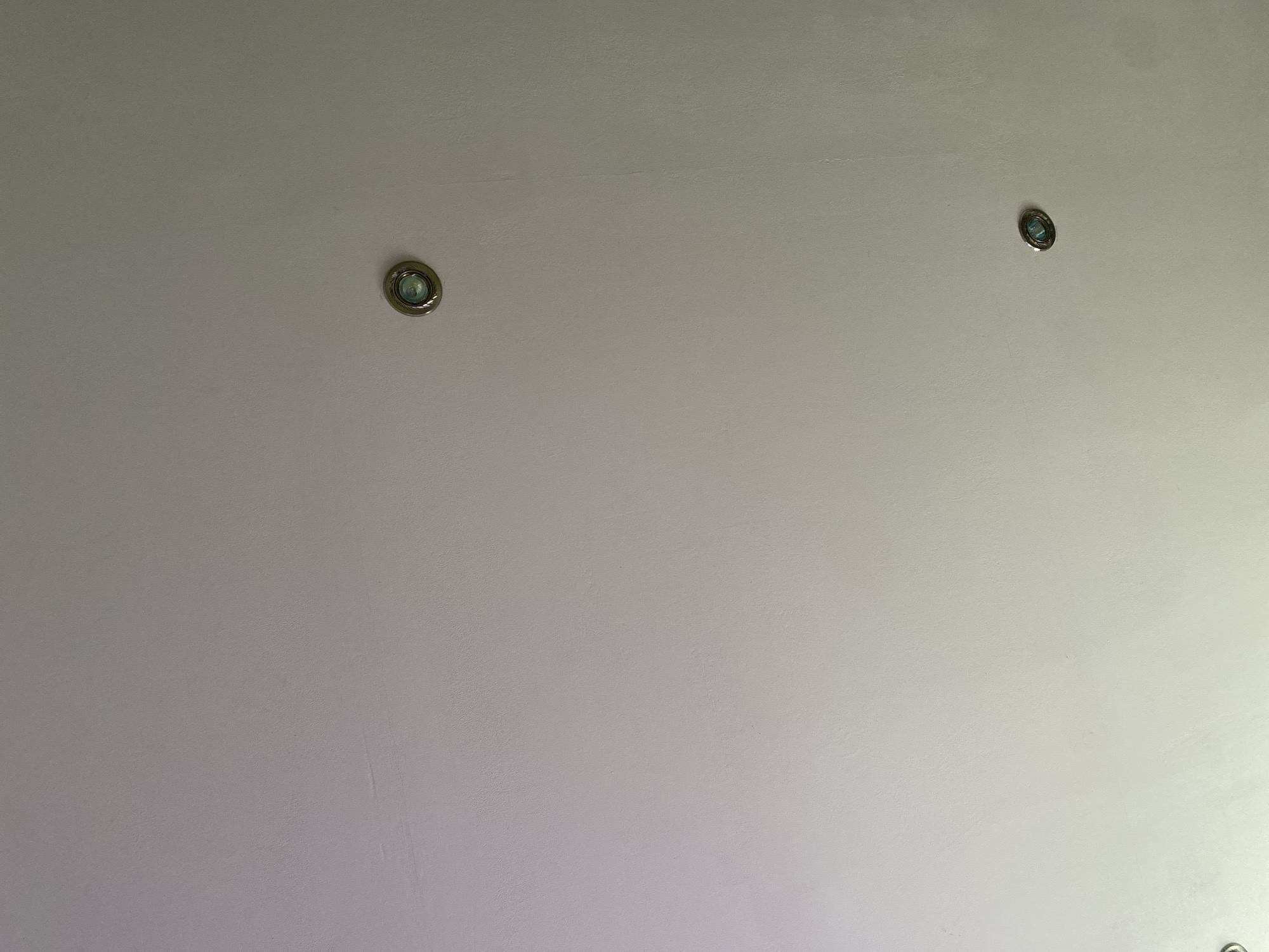 The image shows a white ceiling with three recessed lights. Two of the lights are visible and appear to be working, casting a bluish light. One light is located in the upper right portion of the image, and another is in the lower right. A third light is partially visible in the lower left corner. The ceiling is textured and appears to be made of plaster. There are no people or other objects visible in the image. The location is described as Shiryaevo, Russia, but no specific context for the image is provided.