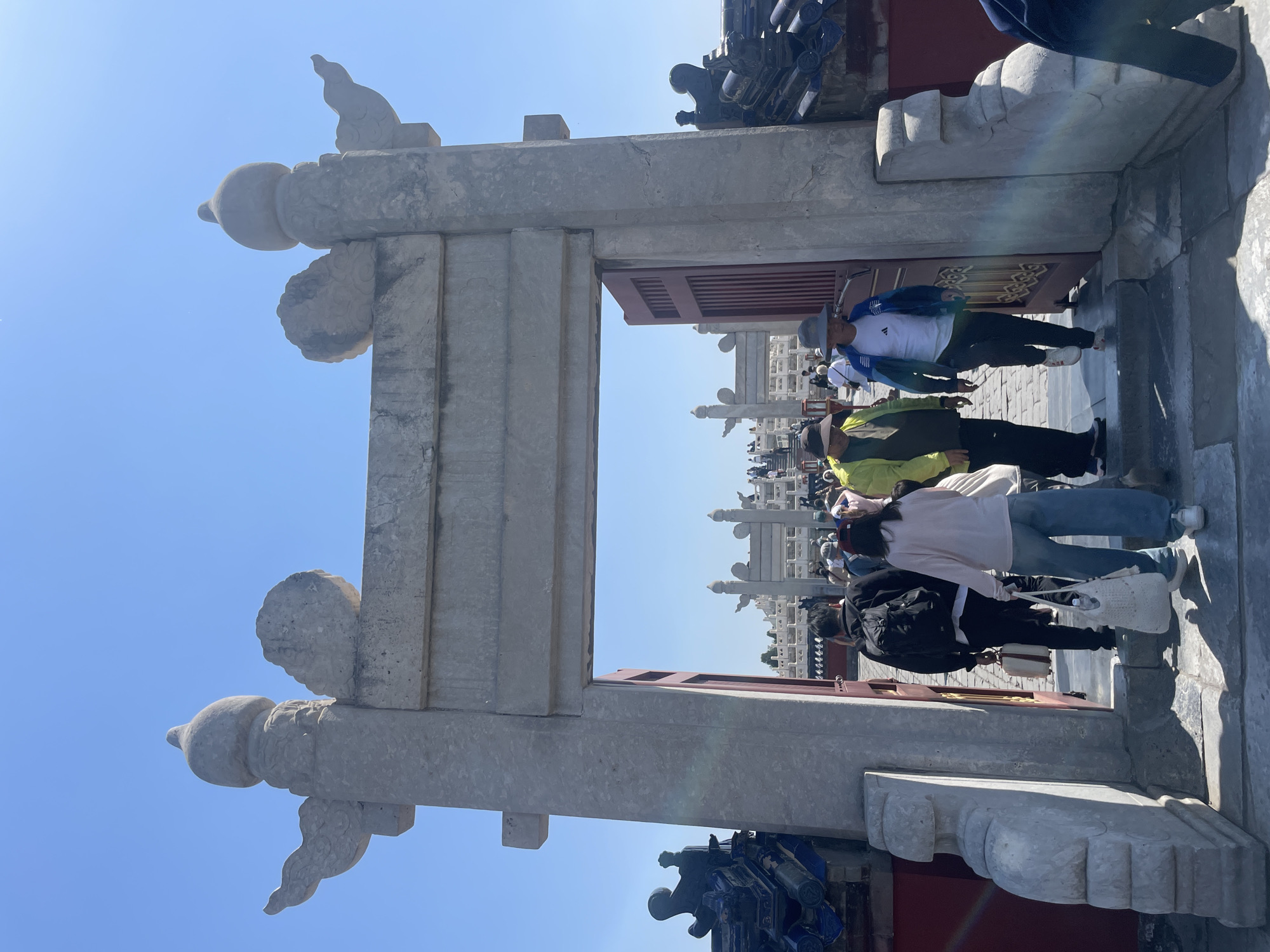 The image depicts a stone gate with ornate carvings, possibly a traditional Chinese structure, in the Dongcheng District of China. Several people are walking through the gate, including a person in a white shirt and blue jacket, a person in a yellow jacket, and a person in a pink shirt. In the background, there is a building with similar carvings and architectural details. The sky is clear and blue.