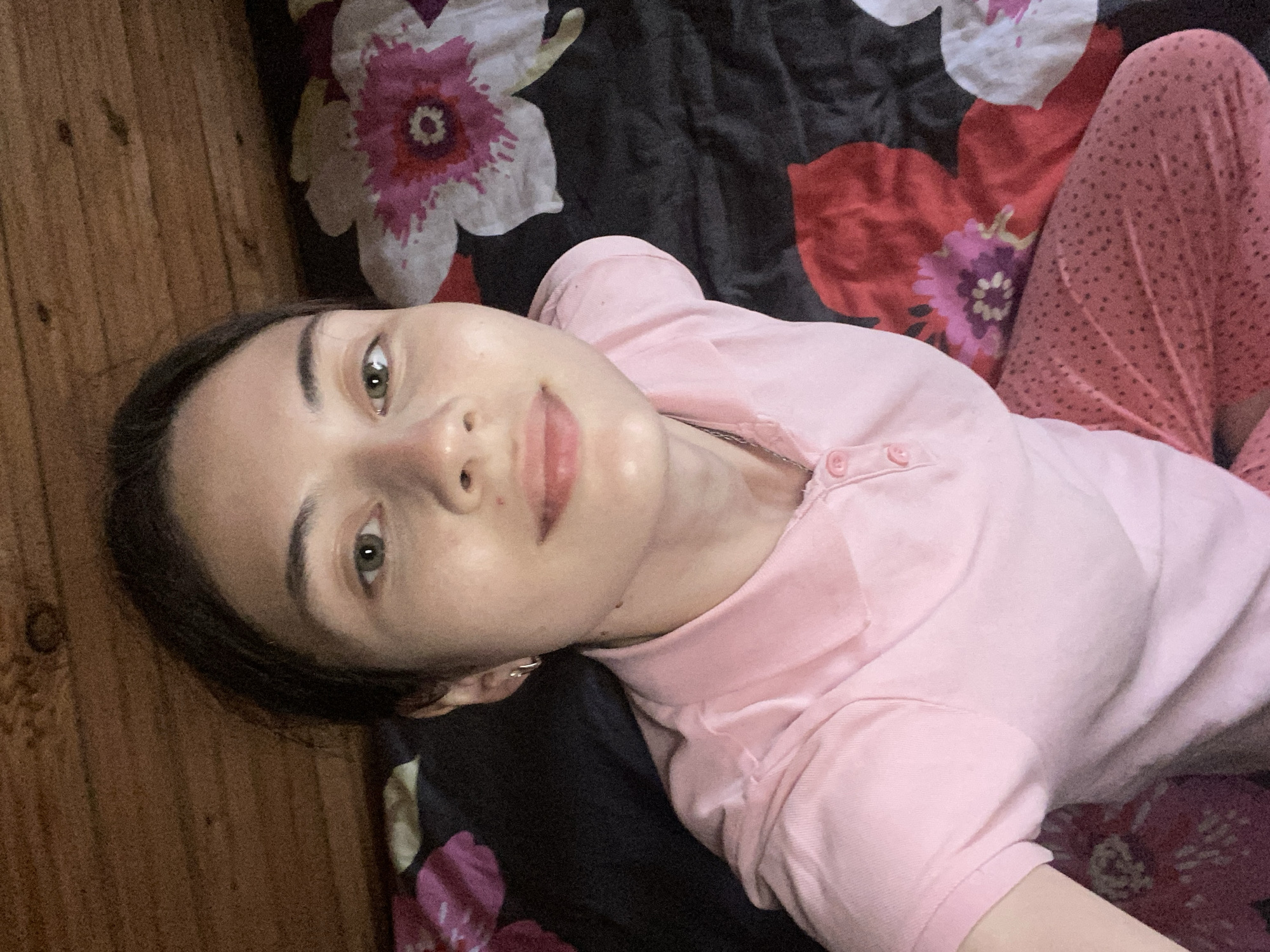 The image shows a young woman lying on a bed with floral patterned sheets. She is wearing a pink polo shirt and pink polka dot pajamas. Her face is looking directly at the camera. The background shows a wooden ceiling. The overall scene is indoors, likely a bedroom. No specific location within Votkinsk, Russia is identifiable from the image itself.