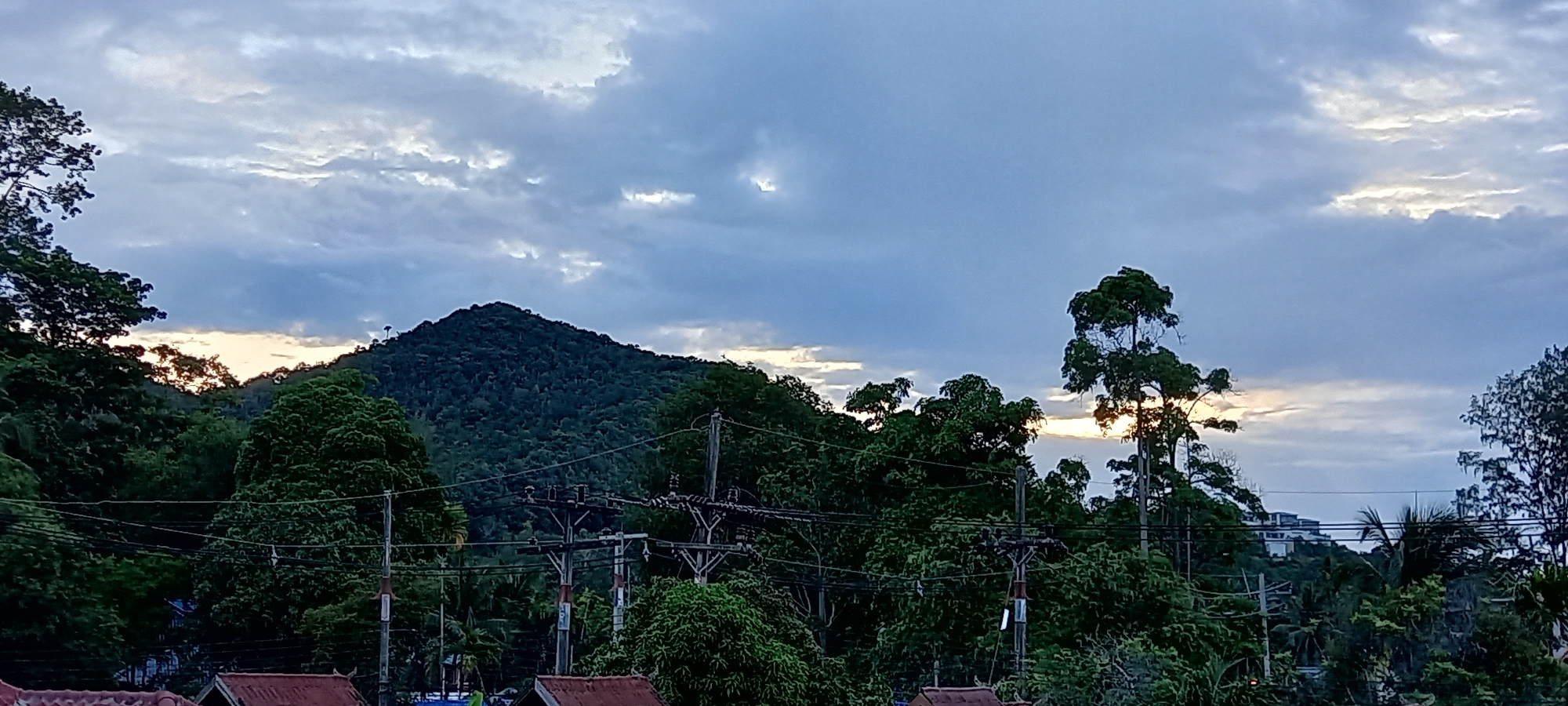 The image shows a mountain covered in lush greenery under a cloudy, pre-dawn sky in Baan Chaloklum, Thailand. A dense layer of trees fills the midground, obscuring the lower portion of the mountain. Power lines intersect the view in the midground. A building is slightly visible to the right of the center. In the foreground are the roofs of buildings.
