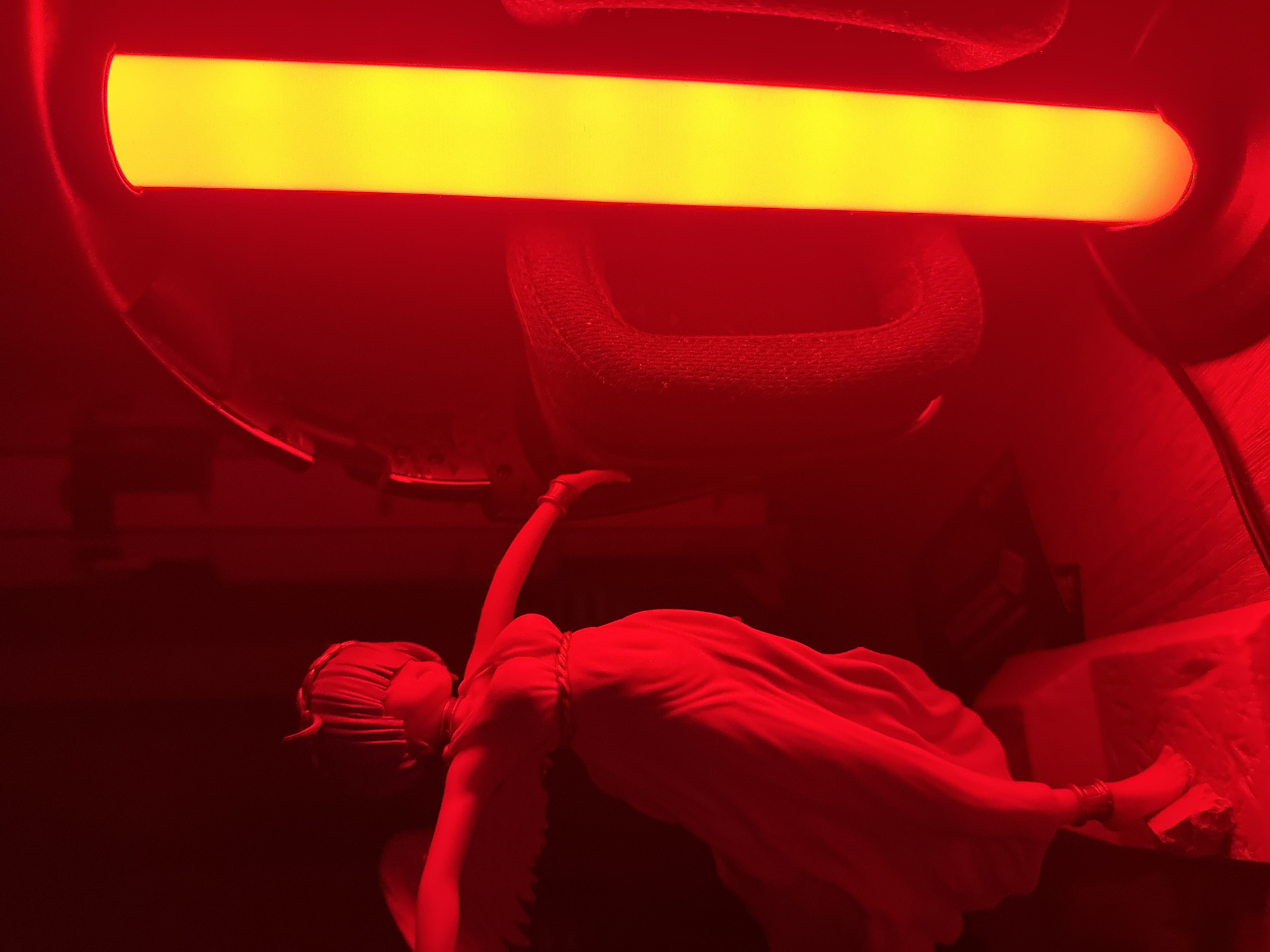 In an indoor scene under red lighting, a statue of a winged figure is positioned to the left. The statue depicts a female figure with wings outstretched and wearing a flowing robe. To the right of the statue, a bright yellow cylindrical light source stands vertically. Behind the statue, a portion of a dark object with textured padding is visible, possibly a headset or chair. The overall environment is dimly lit, with the red ambient light casting shadows and highlighting the yellow glow of the cylindrical light. The scene is static, with no apparent human interaction. The location is Unknown, Unknown.