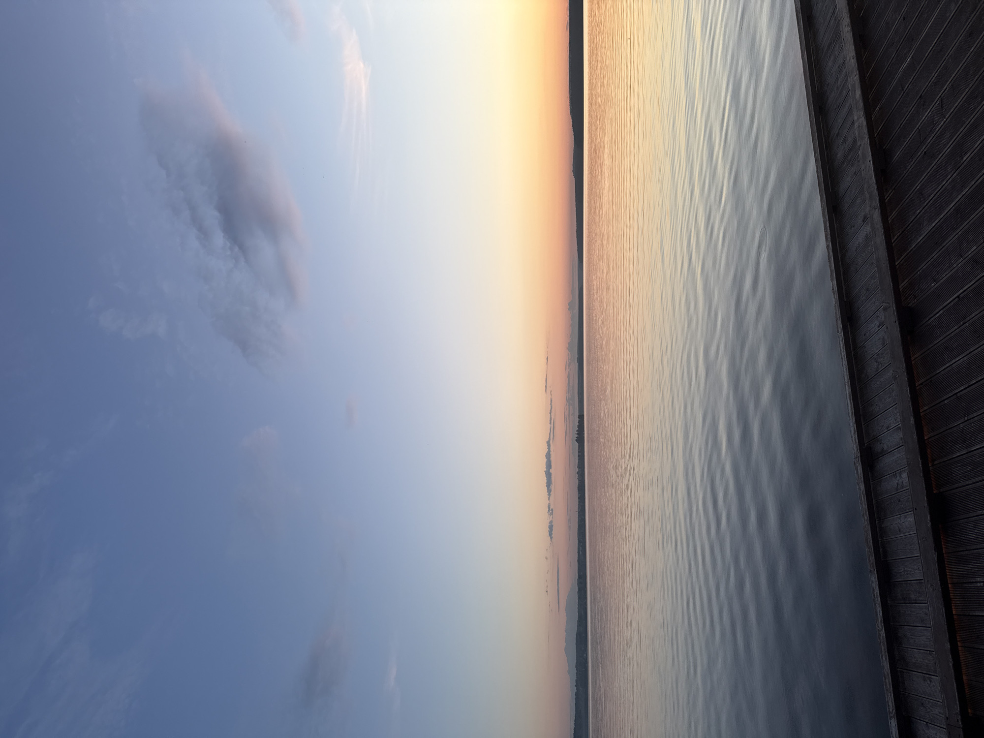 The image presents a serene landscape captured at either sunrise or sunset over a large body of water, identified as the Votkinsk Pond (Votkinsk Reservoir) in the city of Votkinsk, Russia. The sky, occupying the upper two-thirds of the vertical composition, displays a color gradient transitioning from a pale blue at the top, through subtle shades of light blue and pink in the middle, to a warm orange-yellow hue along the distant horizon. Wispy, diffuse clouds are scattered across the blue and lighter pink portions of the sky, with more elongated, dark clouds appearing closer to the horizon line.
A narrow, dark silhouette of a distant landmass or shoreline stretches across the entire horizon, separating the sky from the water, with faint, indistinct details suggesting minimal topography. The extensive surface of the water, covering the lower two-thirds of the image, exhibits a calm, rippled texture. It vividly reflects the colors of the sky, with the warmer orange and pink tones dominant in the middle ground, gradually darkening to deeper blues and grays towards the immediate foreground.
In the bottom right corner, a section of a dark, wooden dock or pier is visible. It is constructed from multiple parallel planks, showing distinct grain and texture, and extends diagonally into the frame. No individuals, animals, vehicles, or overt human activities are depicted. The scene conveys a tranquil, unobstructed view of the natural environment at a transitional time of day.