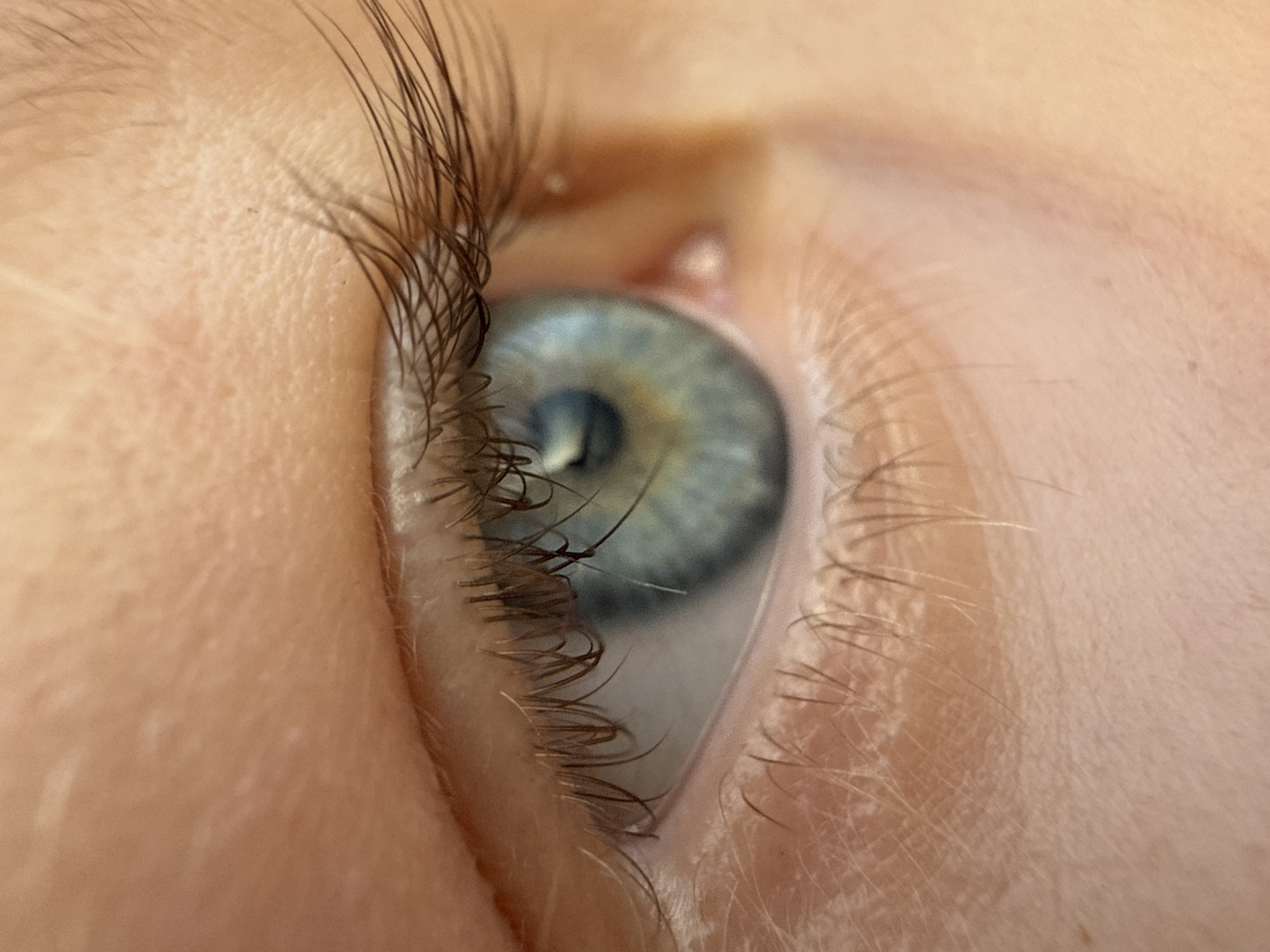 This is a close-up, macro photograph of a human eye, showing intricate details of the iris, pupil, eyelashes, and surrounding skin. The iris displays a blend of blue and green hues, with subtle variations in color and texture. Delicate eyelashes frame the eye, some appearing longer and darker than others. The skin exhibits natural texture, with fine lines and pores visible. The lighting suggests a natural light source, casting soft shadows and highlighting the contours of the face. The image is devoid of any specific urban or natural landscape elements, focusing entirely on the biological subject. There is no information provided in the image that would indicate its location within Baan Hua Thanon, Thailand, or any other geographical area.
