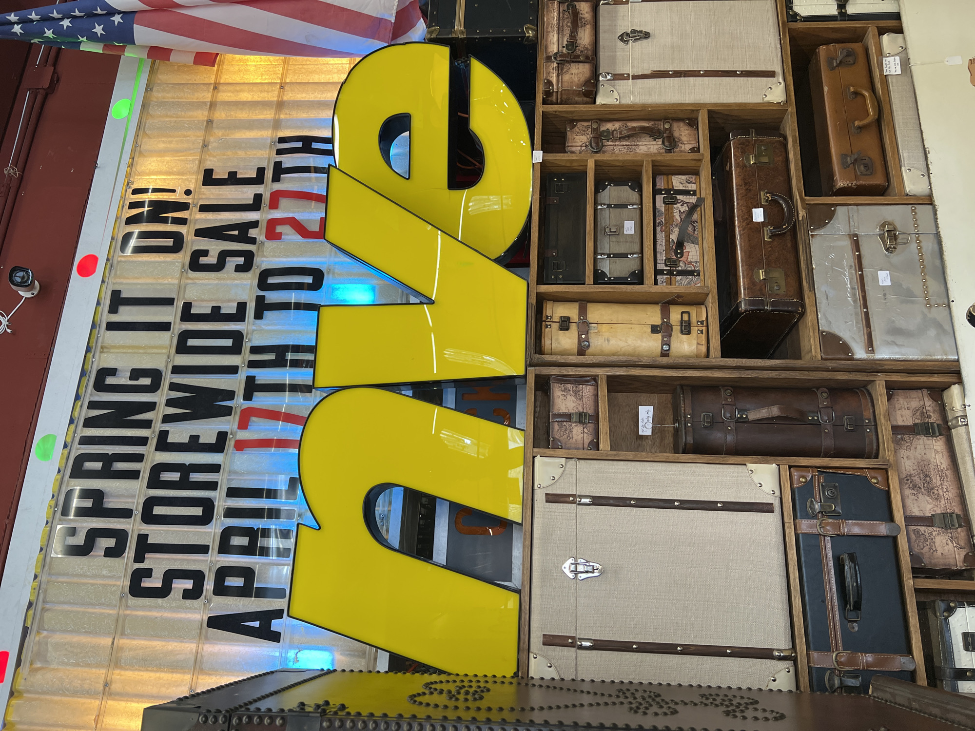The image depicts a storefront in the City of Newburgh, United States of America. A large, yellow "WE" sign dominates the foreground. Behind it, a window display promotes a "SPRING IT ON! STOREWIDE SALE APRIL 17TH TO 27TH." An American flag is partially visible at the top left. To the right of the sign, a wooden shelf unit displays various vintage suitcases and trunks. The setting appears to be an antique or vintage store.