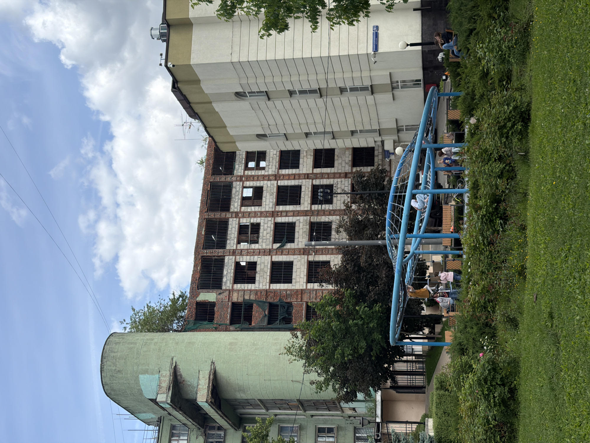 The image shows a cityscape with a playground in the foreground. The playground features a blue climbing structure with several people, including children, using it. To the right of the playground is a green lawn and some bushes.
In the background, there are several buildings. One building is a partially constructed or dilapidated structure with a brick facade and barred windows. Adjacent to it is a more modern building with a lighter-colored facade and rectangular windows. There is also a green cylindrical structure with visible signs of wear and tear.
The sky above is partly cloudy. Street signs are visible, suggesting an urban environment.