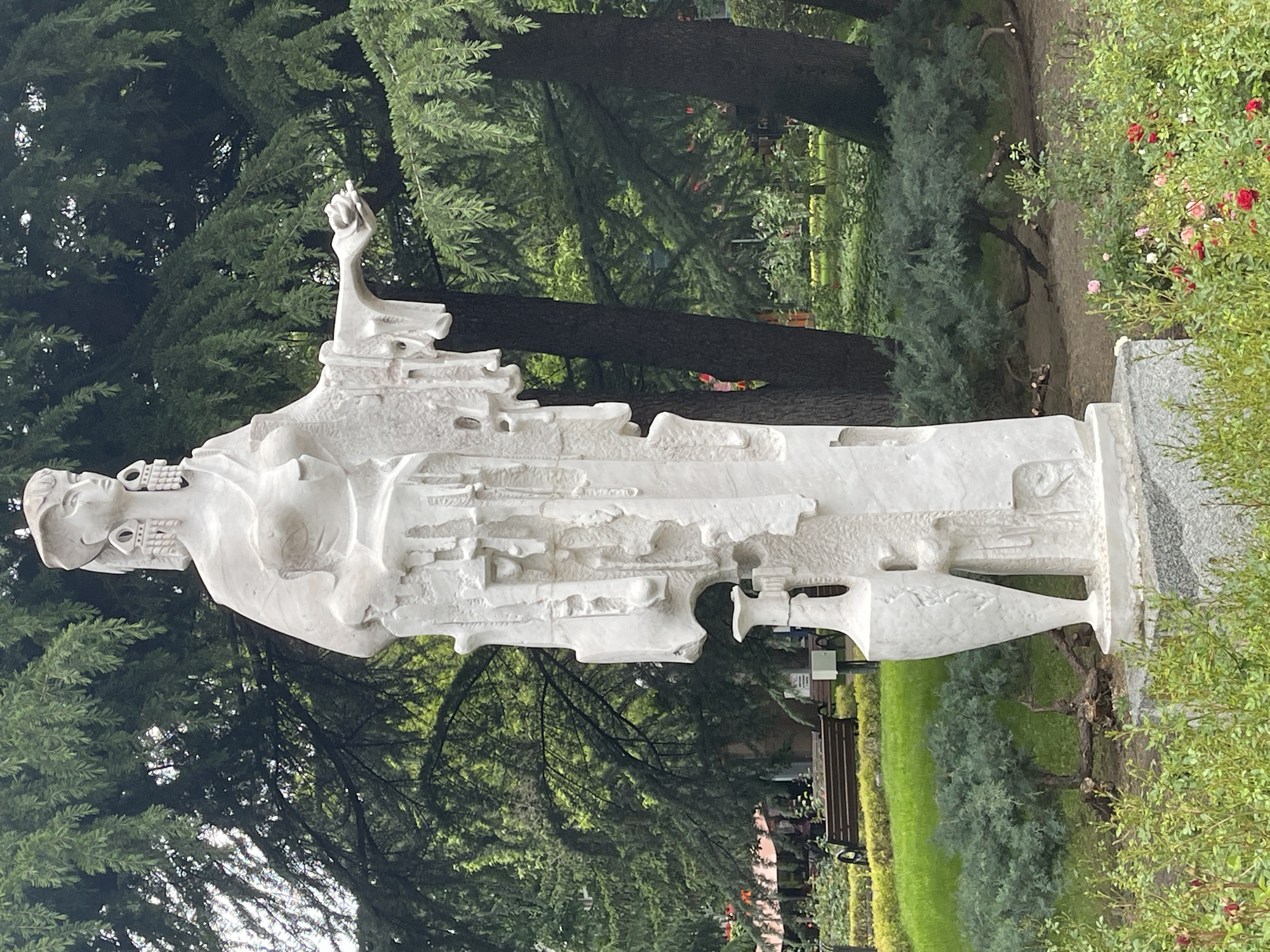 A white, abstract sculpture of a standing figure with its right arm raised stands on a stone base in a park-like setting. The statue is positioned amidst trees and bushes. Red roses are visible in the background. The scene appears to be located in Kazanlak, Bulgaria.
