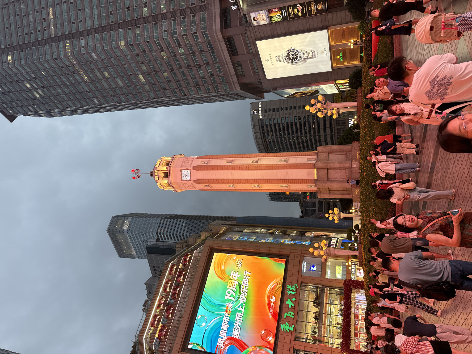 In Yuzhong District, China, a lively pedestrian area is depicted at dusk, illuminated by a mix of artificial lighting and the fading daylight. A prominent clock tower stands as a central focal point, its beige structure adorned with warm, orange light. To the left, a large, vibrant LED screen displays a colorful advertisement. Flanking the clock tower are modern skyscrapers, one to the left with a tiered, illuminated facade and another to the right, a tall, grey building with numerous windows. Below the screen and around the clock tower, a dense crowd of people is gathered, engaged in various activities. Some individuals are walking, others are standing and conversing, and a few are seated. Among the crowd, there are families with children, couples, and individuals. The ground is paved with textured tiles. Lush green foliage and flowering plants are visible in planters surrounding the base of the clock tower and in front of the buildings, adding a touch of nature to the urban landscape. Streetlights with multiple yellow bulbs are strategically placed throughout the scene, casting pools of light. A large billboard featuring a Rolex watch is prominently displayed on the right side of the image, adding a commercial element to the scene. The sky above is overcast, contributing to the dim evening atmosphere.