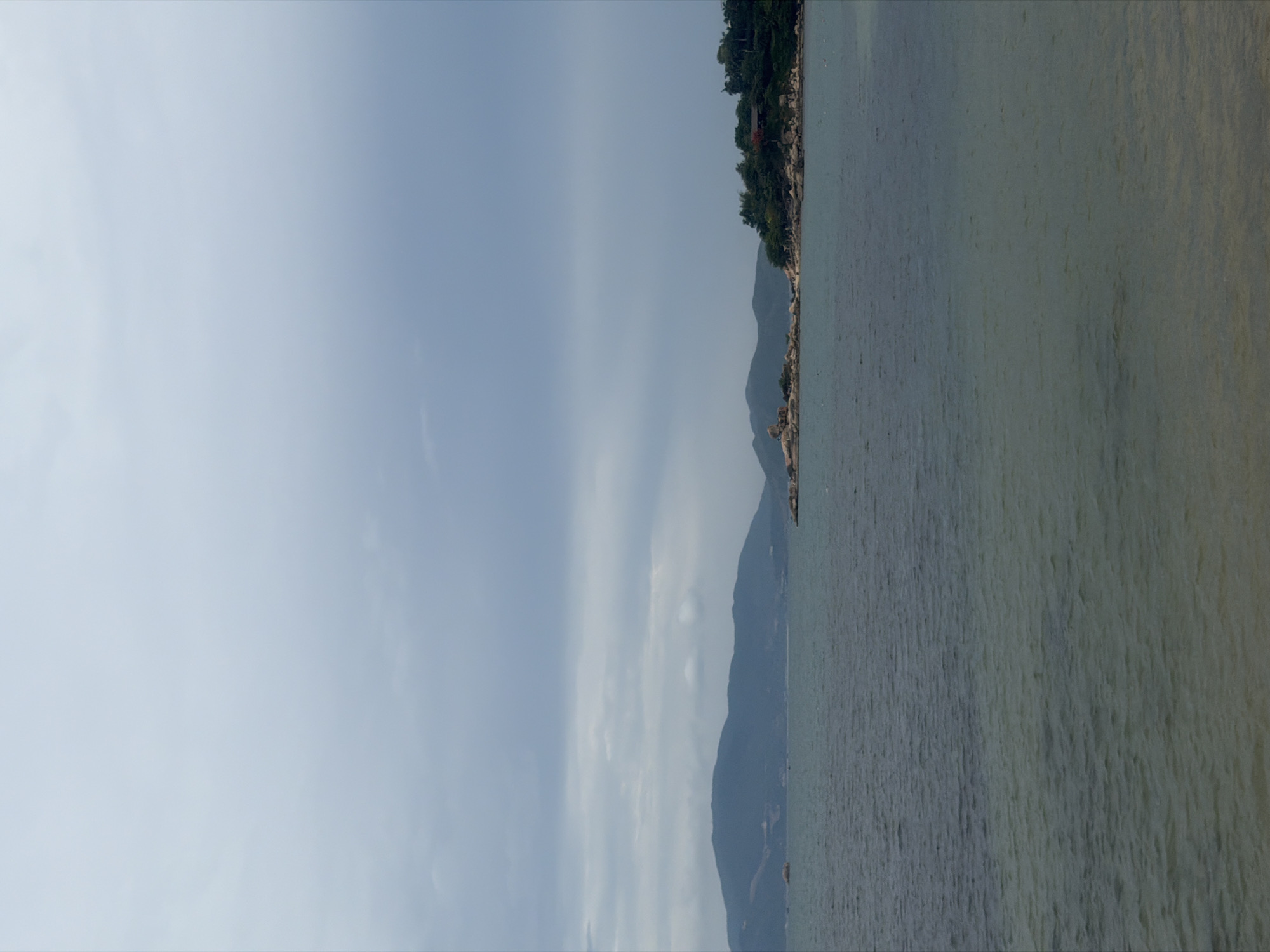 The image captures a view of Bac Nha Trang Ward, Vietnam, featuring a body of water on the right side of the frame. The water appears calm with an even surface texture. On the left side, the sky is visible, showing a uniform, overcast gray color. In the middle of the image, a coastline is present, separating the sky and the water. This coastline features a progression of land masses, potentially hills or mountains, with vegetation visible along the topmost ridge. No people, specific objects, or interactions are discernible in the image.