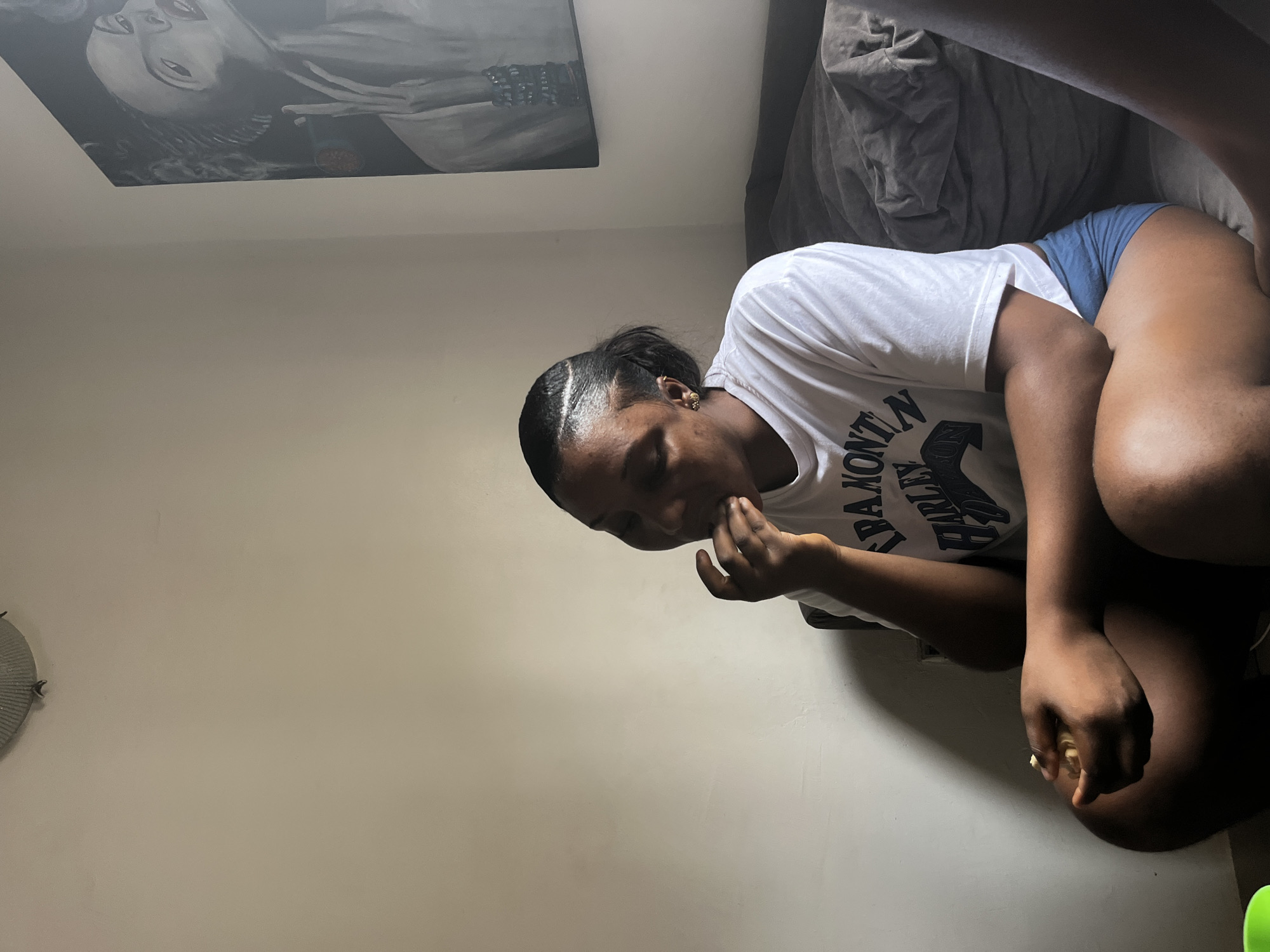 A person is sitting in a room, with a painting on the wall. The person is wearing a white t-shirt with black text and blue shorts. They are holding their hand to their mouth and looking down. The environment appears to be a domestic interior. The city of Abuja, Nigeria is not identifiable from the image.