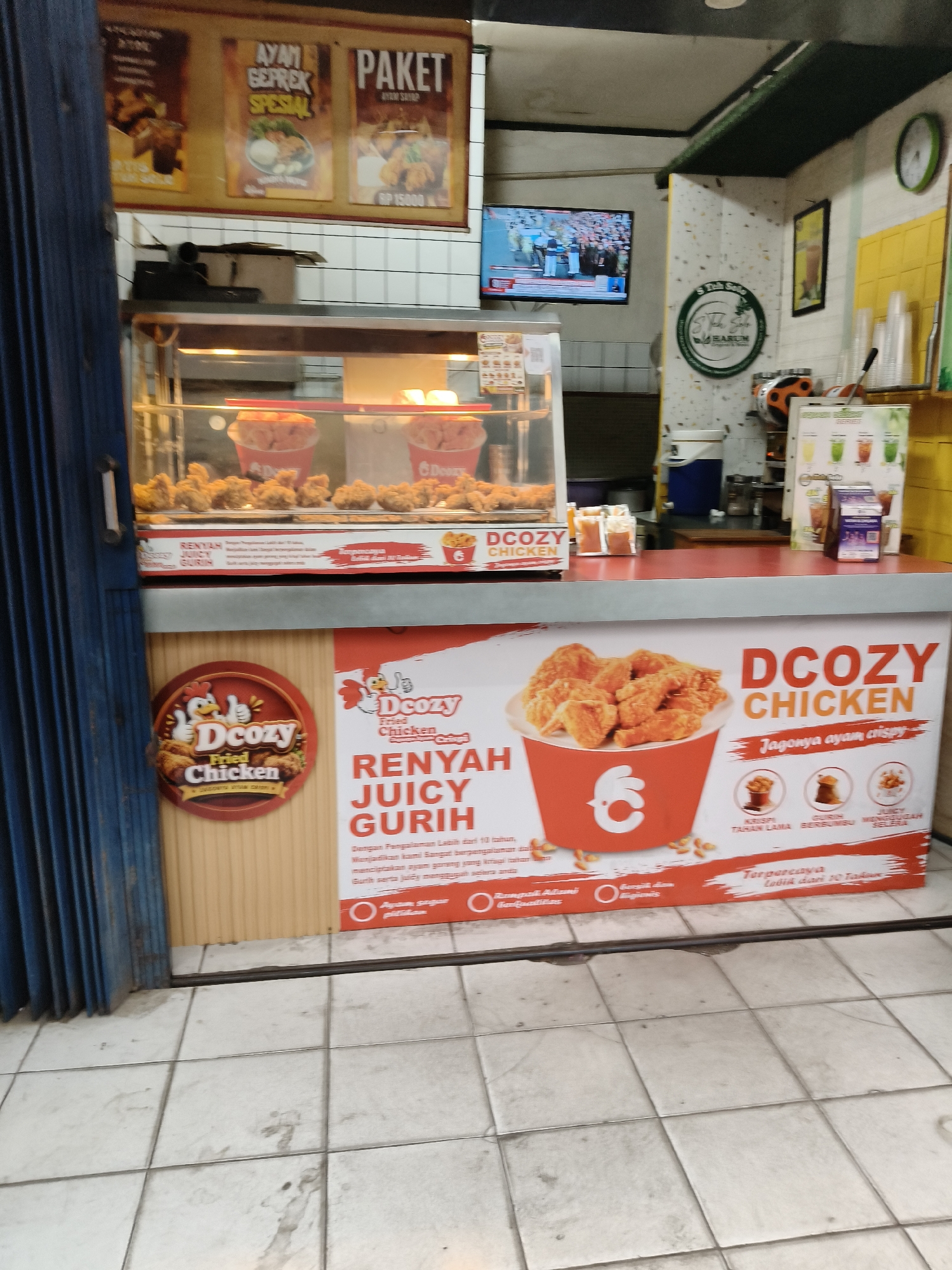 The image shows a street-level outdoor food stall in South Tangerang, Indonesia, likely a fried chicken franchise named "Dcozy Fried Chicken." The stall features a display case filled with fried chicken and buckets of chicken. A large banner on the front of the counter advertises "Dcozy Chicken" with images of chicken and text in Indonesian, highlighting its crispy and juicy qualities. There are also posters on the wall behind the stall advertising menu items like "Paket Ayam Goreng Spesial." A television screen on the wall displays what appears to be a news broadcast. The overall environment suggests a bustling commercial area.