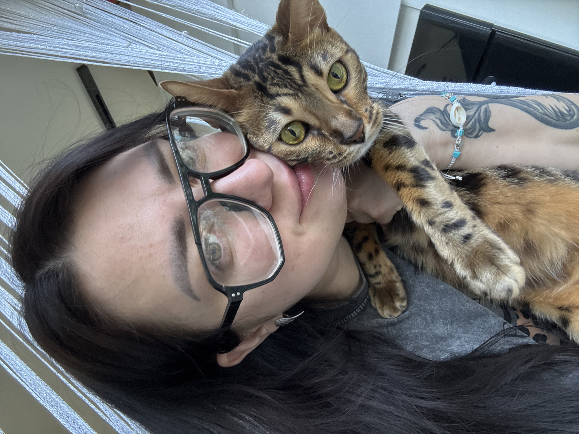 A close-up, eye-level shot captures a person and a Bengal cat in an intimate, affectionate pose, likely indoors. The person, with long dark hair, is lying down, wearing black-rimmed eyeglasses that frame their visible eye. Their face is partially obscured by the cat's head. The Bengal cat, with striking green eyes and distinctive spotted and striped fur, rests its head gently on the person's cheek and chin, its tongue slightly visible.
The person's left arm, visible in the upper right, sports an intricate dark tattoo with leafy or flowing patterns, and a light blue and white bracelet featuring a large cowrie shell and smaller turquoise-colored beads. They appear to be wearing a grey top. In the background, thin, light-colored horizontal lines suggest a woven structure like a hammock or blinds, with a dark rectangular object visible behind the person's head. The scene conveys a moment of calm and close companionship between the person and their pet. No text is visible.