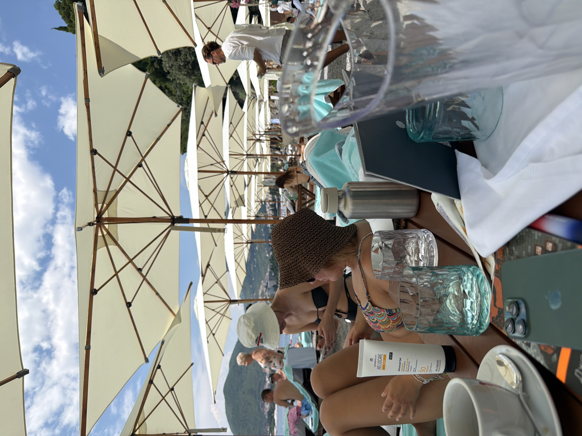 The image captures a vibrant scene at a beach resort, likely in Budva, Montenegro, under a bright, sunny sky with scattered clouds. The setting is characterized by numerous large, cream-colored umbrellas providing ample shade over a row of tables. In the foreground, a woman wearing a colorful patterned bikini top and a woven sun hat is seated at a table. She is holding a tube of sunscreen, visible with the brand name "HELIOCARE" and "Advanced Gel SPF 50" clearly printed on it. Beside her are two textured glass tumblers, one appearing to contain ice or condensation.
In the background, several other individuals are visible, some seated at tables, others milling about. One man in a white shirt is standing and looking towards the tables. The arrangement of umbrellas suggests a well-established beach club or restaurant. The overall atmosphere appears relaxed and leisurely, typical of a summer day at a popular tourist destination. The presence of umbrellas, beachgoers, and the implied proximity to the sea point towards a vacation or leisure activity. The lighting indicates it is daytime, likely late morning or afternoon. The background reveals a glimpse of lush green hills and possibly a town, consistent with a coastal Mediterranean location like Budva.