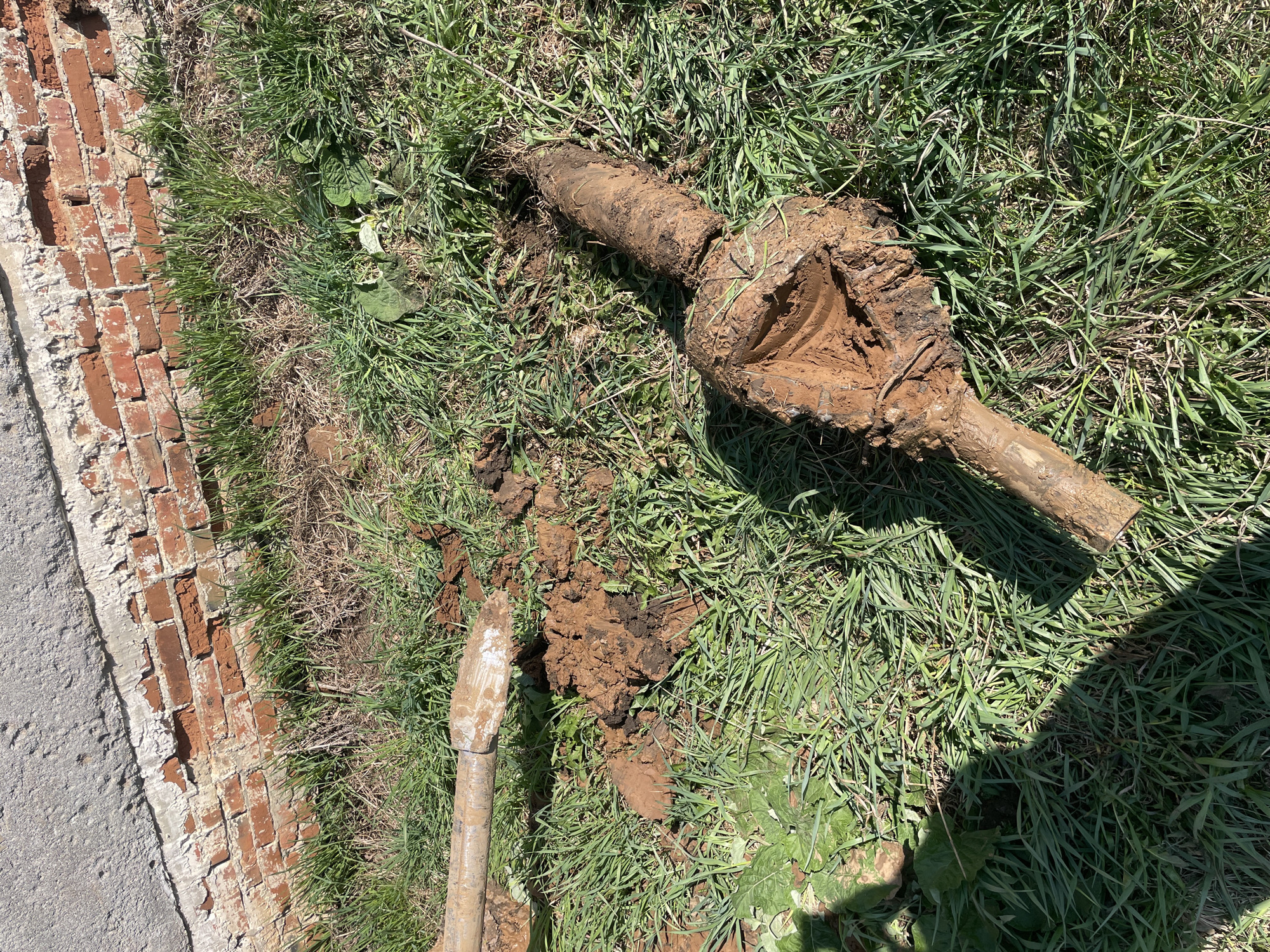 The image depicts a partially excavated pipe fixture coated in mud, resting on grass and dirt. A brick wall runs along the left edge of the frame, bordered by a section of concrete. A shovel is partially visible, also covered in dirt, near the excavated area. There are no people visible.