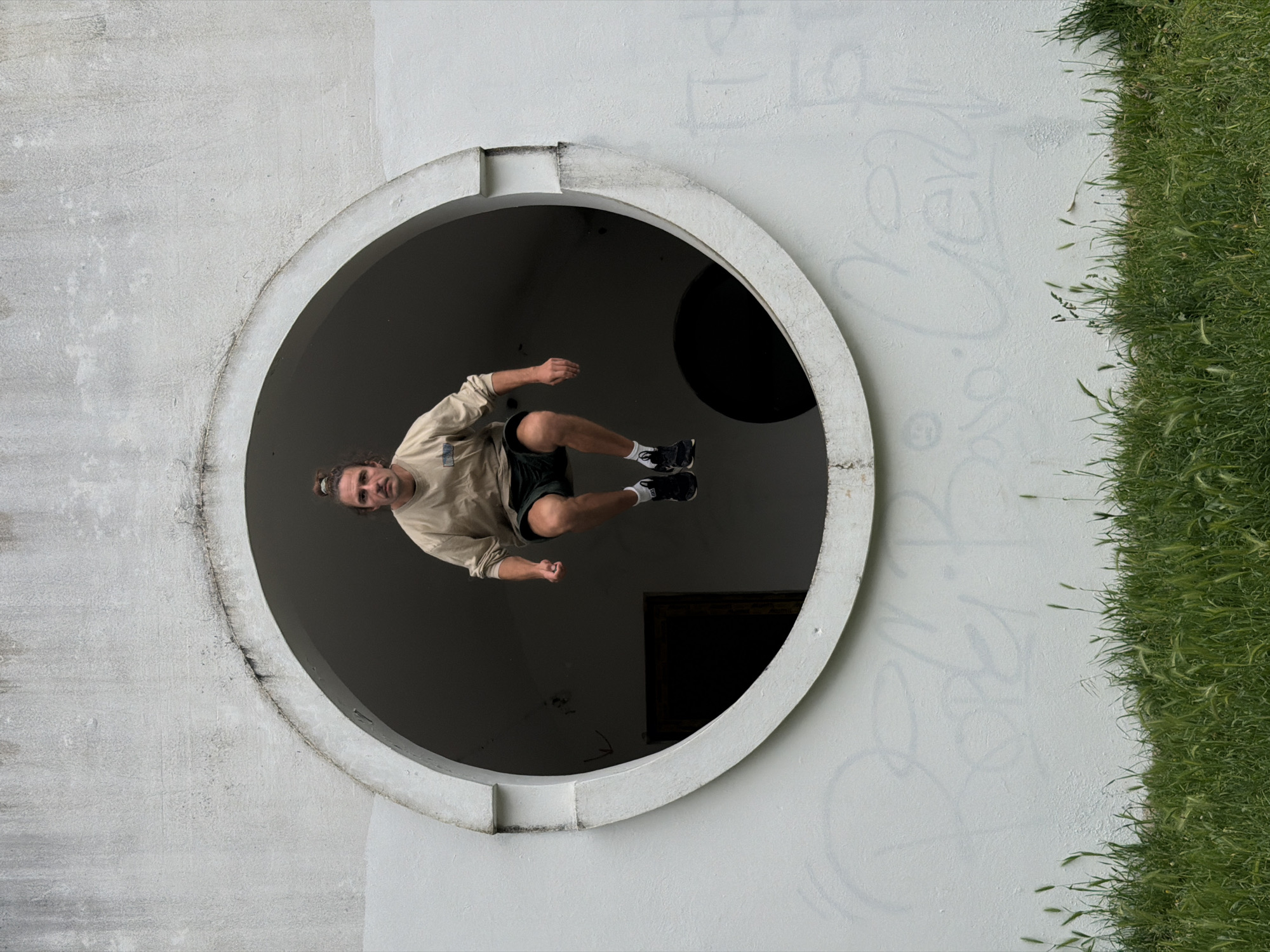 A low-angle shot shows a man executing a jump through a circular opening in a white concrete structure in Petrovac centar, Montenegro. The man has a tan complexion and light brown hair pulled back in a bun; he wears a light tan long-sleeved shirt, dark shorts, white socks, and black and white sneakers. His body is positioned horizontally within the circle, as if in mid-air.
The circular opening is part of a larger white concrete wall, which shows signs of weathering and faint graffiti. To the right of the wall is a dense patch of green grass. Inside the opening, the background is dark and features an unidentifiable black object and a dark rectangle, likely a door or window.