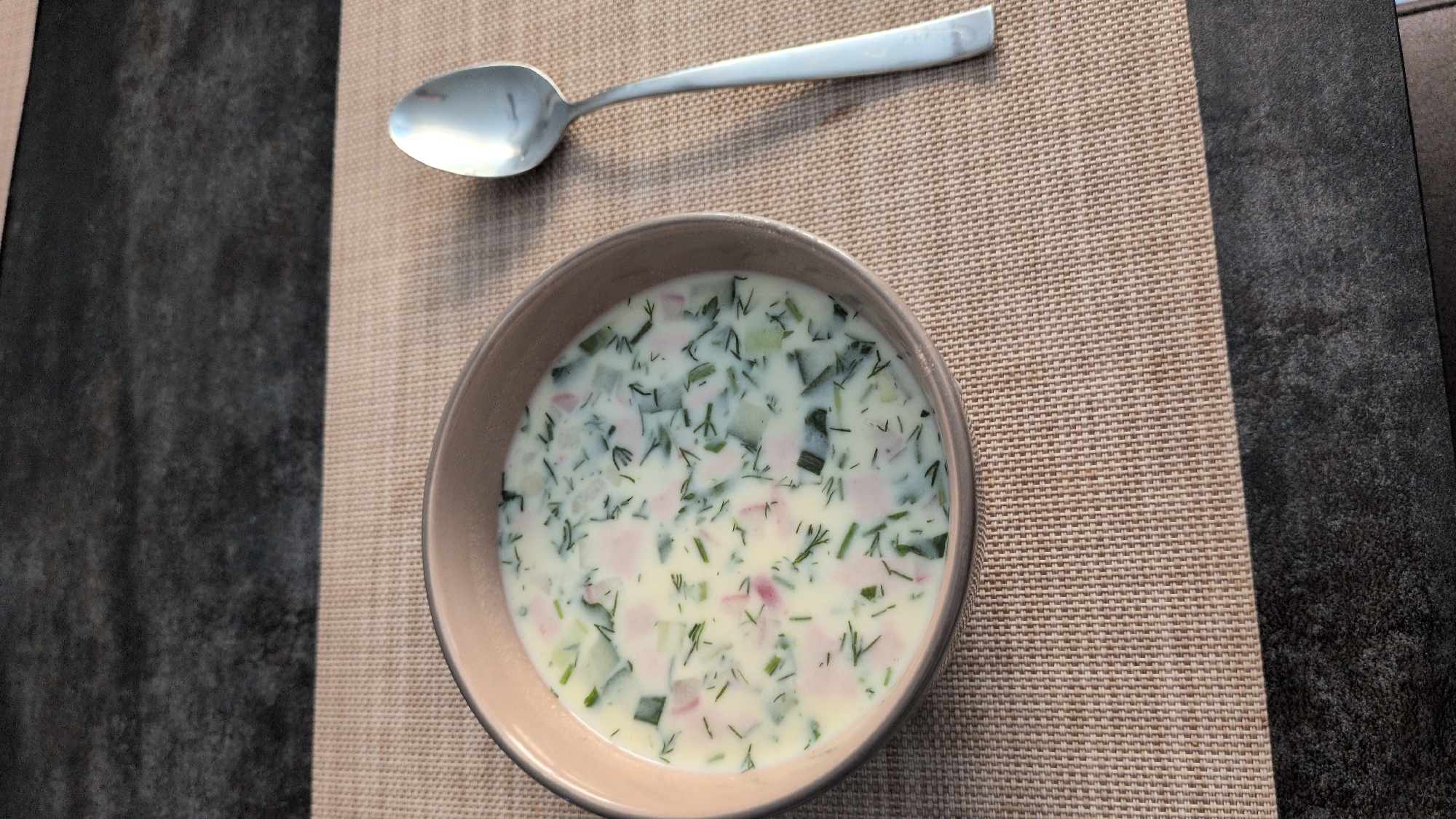 A close-up, overhead shot shows a bowl of cold soup, likely Okroshka, a traditional Russian dish, placed on a woven placemat. The soup is a pale white liquid with finely diced green cucumbers, pink pieces of ham or sausage, and green herbs, possibly dill. A silver spoon rests on the placemat above and slightly to the left of the bowl. The placemat is a light beige color with a woven texture. To the right of the placemat, a dark gray, textured surface, possibly a tabletop, is visible. The lighting appears to be natural and even, suggesting daytime. No people are visible in the image. The scene is simple and focused on the food and dining setup. There is no visible text in the image. The setting is domestic, likely a dining table.