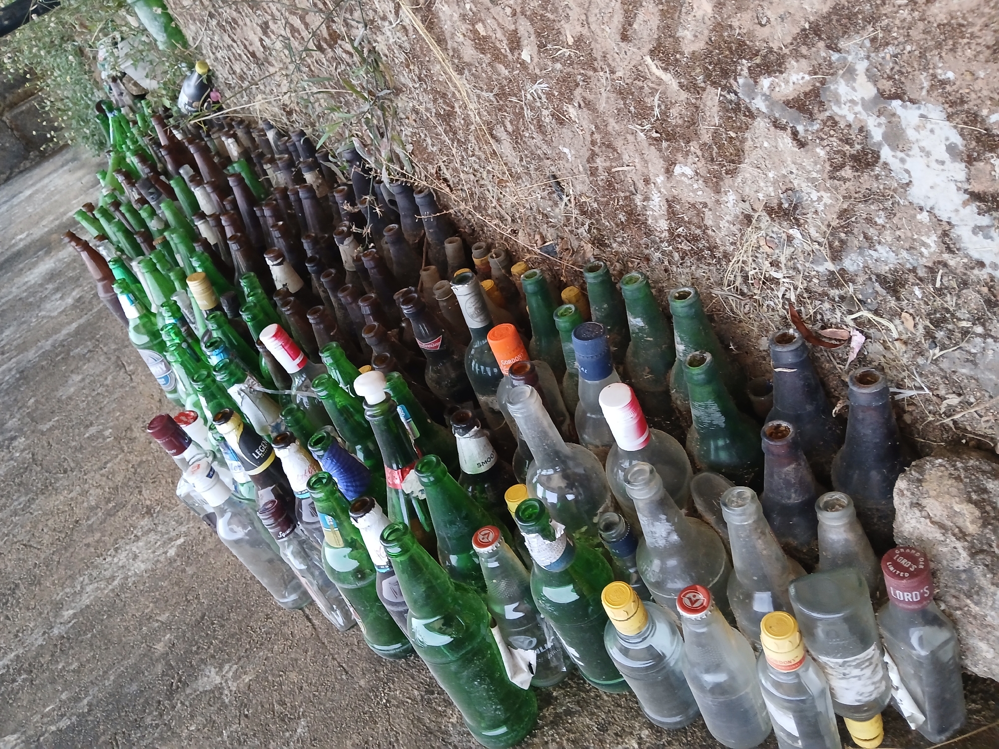 The image shows a large collection of empty glass bottles, mostly beer and liquor bottles, discarded alongside a rough, possibly concrete, surface and against a dirt or rock wall. The bottles are of various sizes, shapes, and colors, with a predominance of green and brown. The scene appears outdoors, with sunlight illuminating the area. While a city is mentioned, there's no evidence in the image to pinpoint the location as Jos, Nigeria; it simply shows discarded bottles in an outdoor environment.