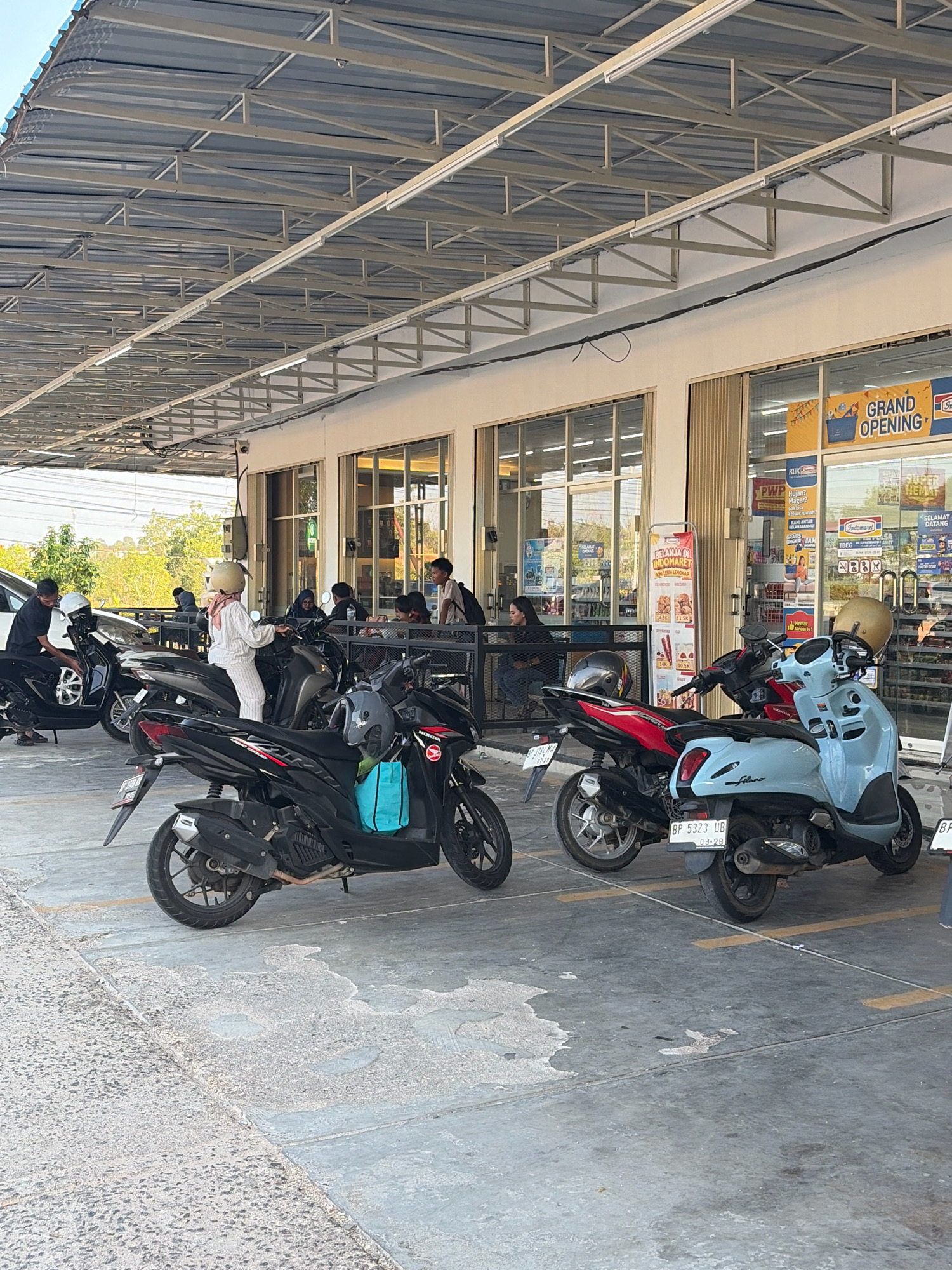 The image depicts an outdoor area in front of a row of shops, likely a convenience store or minimarket, in Tanjung Pinang, Indonesia. A grey metal awning structure with visible support beams and fluorescent lighting covers the walkway and parking area. Parked scooters and motorcycles are prominently featured, with at least three visible in the foreground. One light blue scooter has a license plate that reads "BP 5323 UB". A black motorcycle to its left has a license plate that is partially visible. A black motorcycle is also seen parked further left, with a man in a dark shirt adjusting it.
Several people are visible. A woman in a white outfit and helmet is seated on a scooter in the middle ground. Other individuals are seated at tables or standing near the entrance of the shops, which have large glass windows. A banner inside one of the shops announces "GRAND OPENING". The ground is paved with concrete, with yellow parking lines visible. Trees and foliage are visible in the background. The scene appears to be during daylight hours, with the sun casting shadows.
