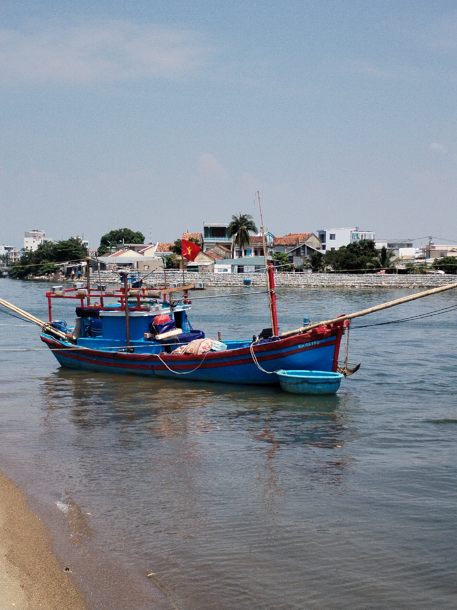 A blue and red fishing boat is anchored near a sandy shore in Nha Trang Ward, Vietnam, under a clear sky. The boat features a red Vietnamese flag, ropes, and a small blue tub hanging off the front. The water is calm with slight ripples, reflecting the boat and sky. The shore consists of light brown sand. In the background, a cityscape includes buildings, trees, and a seawall. The overall scene is a tranquil coastal setting.