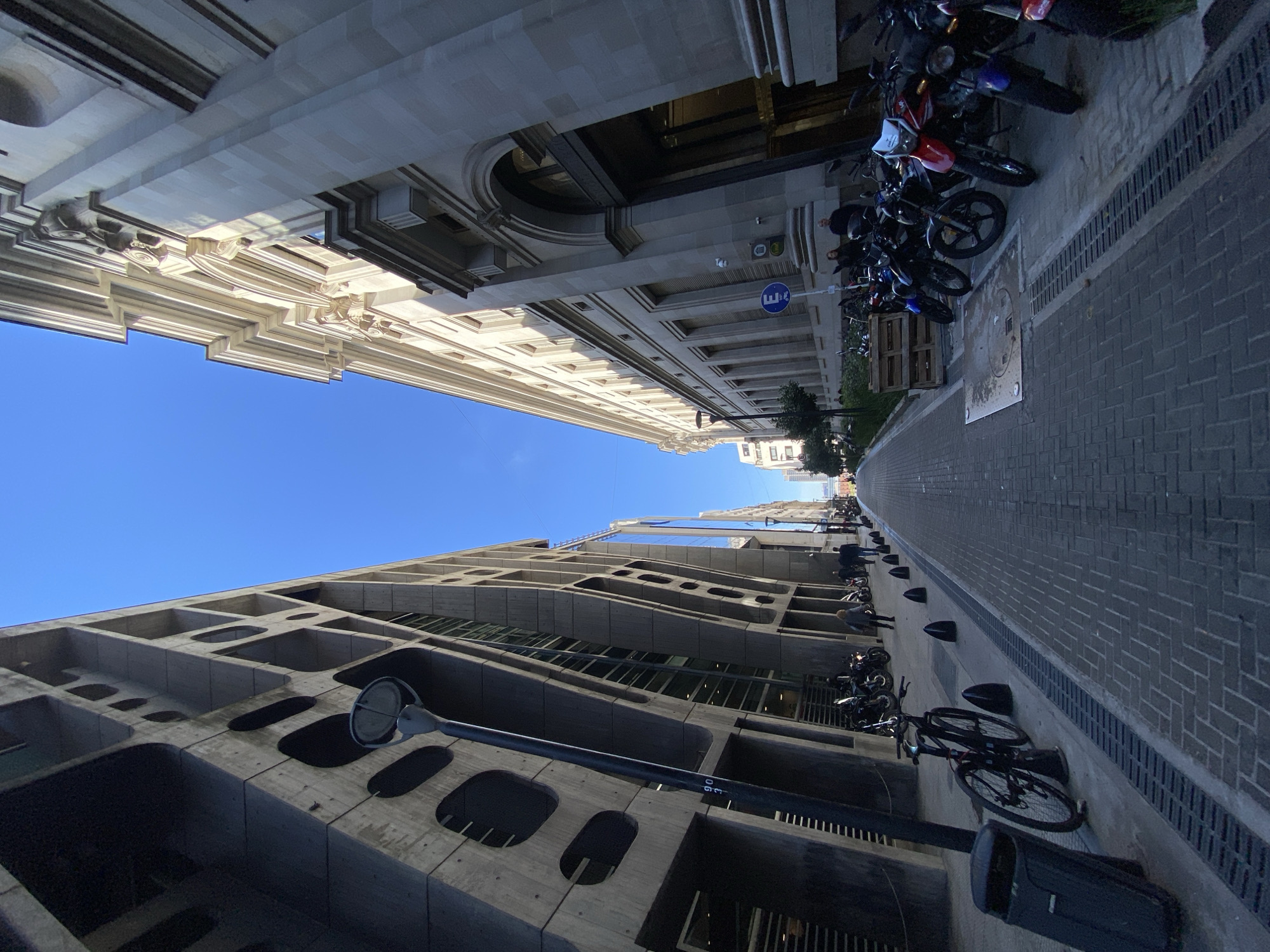 The image shows a street scene in Buenos Aires, Argentina, specifically looking up between two buildings. The building on the right is a classic, ornate style structure, while the building on the left is a modern, concrete building with a grid-like facade. The sky is clear and blue.
Along the right side of the street, a row of motorcycles is parked. There are bicycles parked along the left side of the street. The street is paved with cobblestones and there is a narrow sidewalk on either side. Some distant figures of people can be observed.