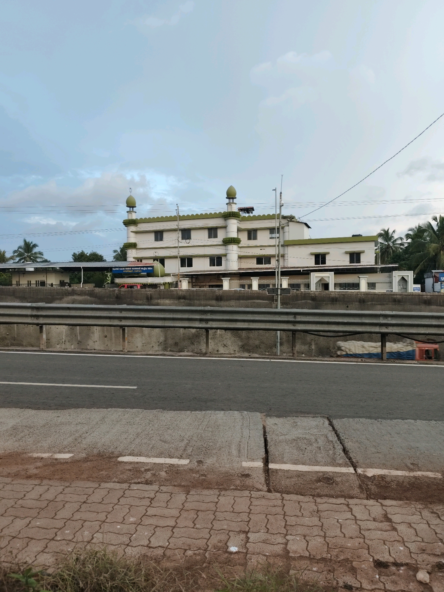 The image depicts a mosque in Kuttippuram, India, viewed from across a road. The mosque is a multi-story building with white walls and green trim, featuring multiple domes. A highway guardrail runs across the foreground, separating the mosque from the road. The road itself is paved, with visible lane markings and a concrete barrier. Palm trees are visible in the background, suggesting a tropical climate. The sky is partly cloudy, with a mix of blue and grey. The overall scene is a typical roadside view of a religious structure in an urban or suburban environment.