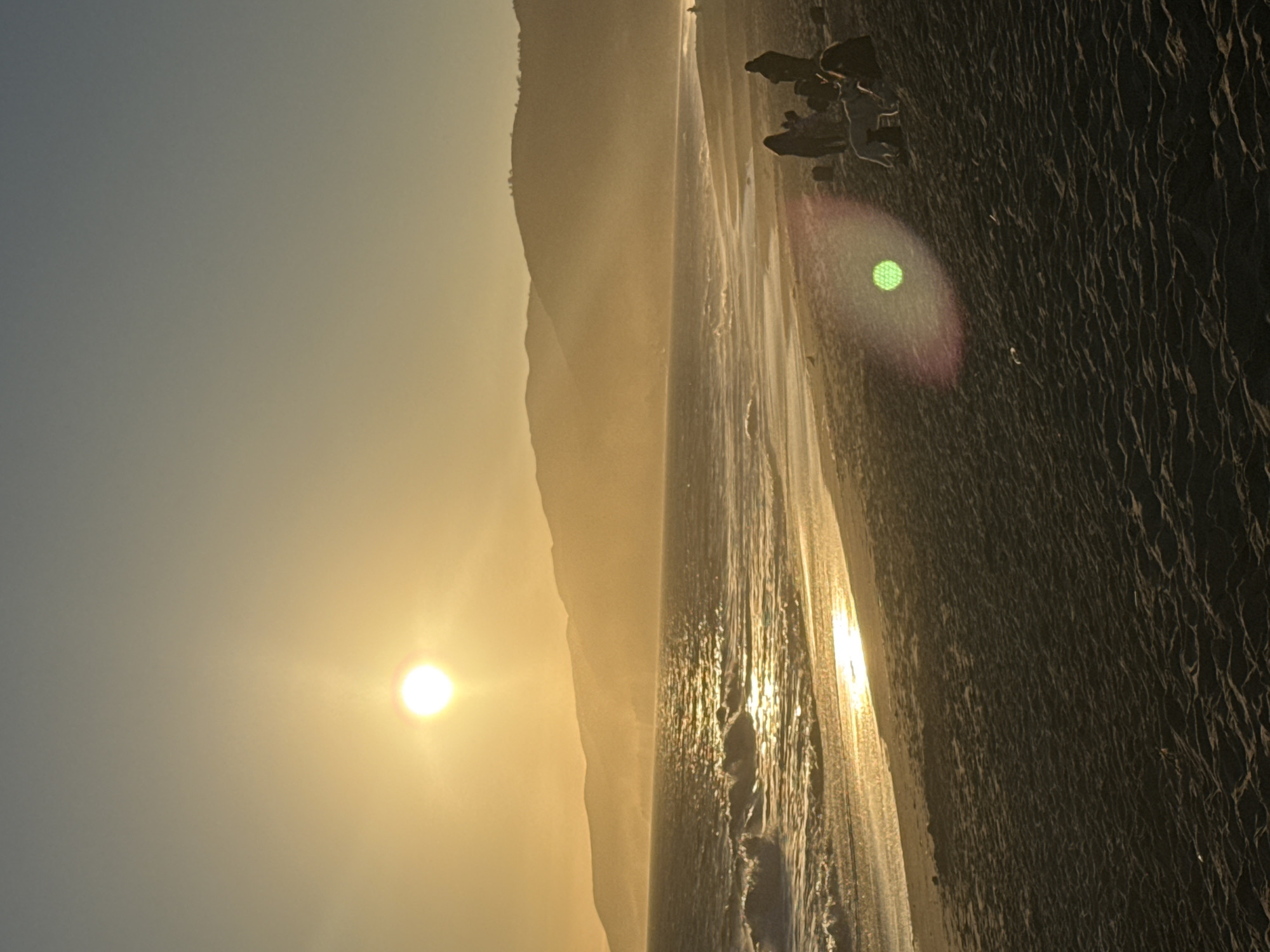 This is a vertical, medium-shot, eye-level, natural light photograph of a beach in Malibu, California, USA, during sunset. The sun, a bright orb, is positioned in the upper left quadrant of the frame, casting a golden glow over the scene. The sky transitions from a clear, pale blue at the top to a warm yellow-orange gradient near the horizon. Distant mountains form a silhouetted backdrop, shrouded in a light haze. The ocean is calm, with gentle waves breaking on the shore, their surfaces shimmering with the sun's reflection. The wet sand near the water's edge reflects the golden light, creating a bright band along the shoreline. Further up the beach, the dry sand is textured with small ripples.
In the middle ground, on the right side of the frame, a group of people are gathered. One person appears to be standing, while others are seated or reclining on the sand. A white dog stands near the seated figures. To the left of this group, a bright green lens flare with a pink halo is visible, also on the sand. The overall atmosphere is tranquil and serene, characteristic of a sunset on the California coast.