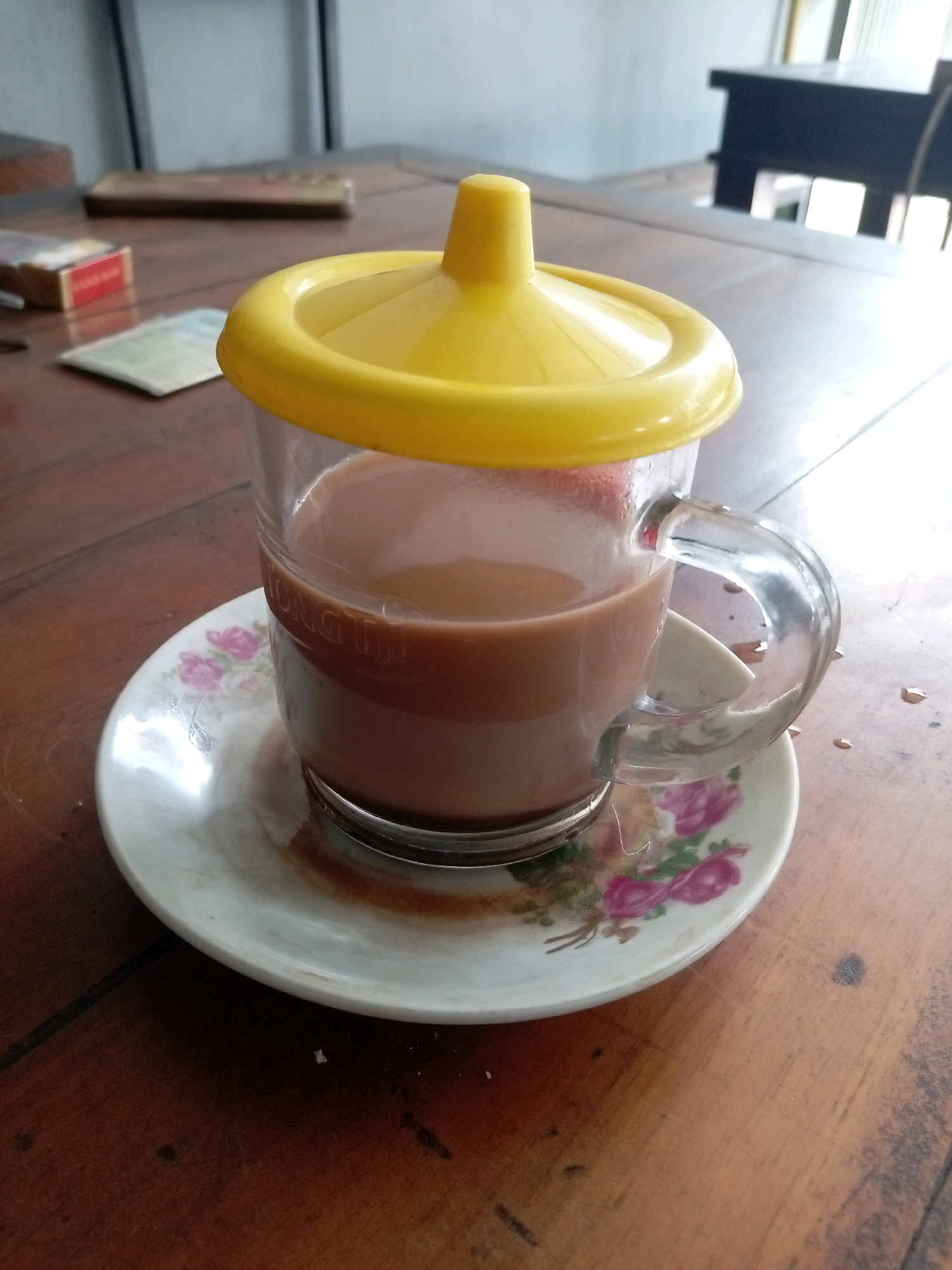 A close-up, slightly angled shot shows a clear glass mug filled with a light brown beverage, likely coffee or chocolate milk, sitting on a saucer with a floral pattern of pink and green flowers. A bright yellow lid with a raised knob is placed on top of the mug. The mug has a glass handle and text is visible around its rim, though it is mostly obscured by the drink. The saucer is slightly stained, with a few small drops of liquid on the wooden table surrounding it. In the background, out of focus, are a red and gold cigarette pack on the left, a wooden object behind it, and a glimpse of a dark wooden table and chairs, suggesting an indoor setting. The lighting appears to be natural, coming from the right, creating highlights on the glass and saucer. The overall scene is casual and appears to be set in a home or a small eatery. This image was taken in Mojokerto, Indonesia.