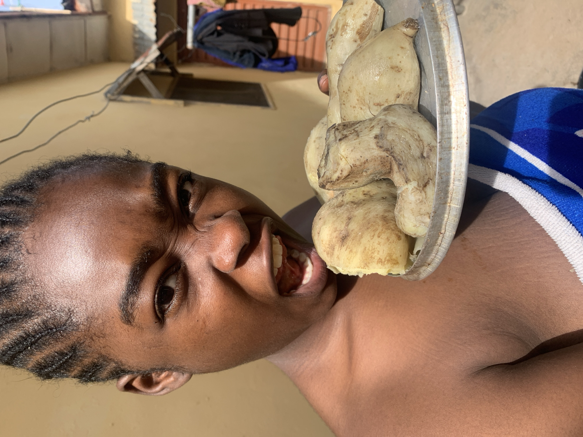 A close-up shot captures a young person with braided hair, a bare chest, and a blue and white striped towel wrapped around their lower body, holding a metal plate with what appear to be boiled yams. The person's face is contorted in a grimace, with their mouth open wide as if about to bite into a yam. The background shows a yellowish building with a corrugated metal roof and laundry hanging on a line. The scene suggests an outdoor setting, possibly in Rafin Par, Nigeria, given the architecture and clothing styles. The lighting indicates it is daytime.