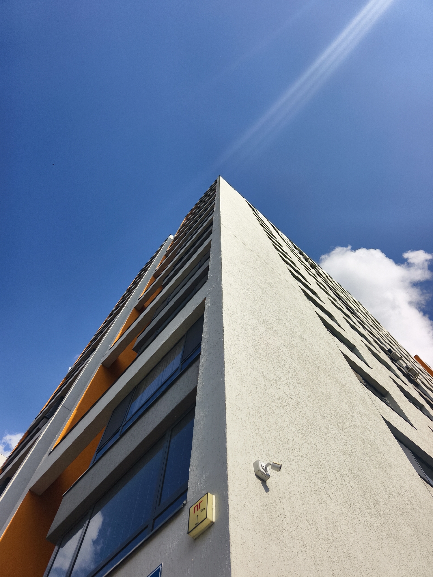 An extreme low-angle photograph captures a modern multi-story residential building located in Tyumen, Russia, viewed upwards along its prominent corner. The building exhibits a light-textured, off-white facade on its right face and a contrasting dark grey facade with intermittent bright orange horizontal accents on its left face. Dark-framed windows are visible across both facades, reflecting the predominantly clear, vibrant blue sky and a solitary white cumulus cloud in the upper right quadrant. A white, bullet-style CCTV camera is affixed to the light-colored facade, casting a distinct shadow. Below the camera, a small rectangular yellow sign bearing the red Cyrillic characters "ПГ" and an upward arrow is mounted. Multiple white exterior air conditioning units are present on the upper levels of the building's right facade. Strong direct sunlight produces two distinct parallel lens flares across the upper-center sky. A small, dark avian form is discernible in the upper-left sky. No human figures are present within the frame.