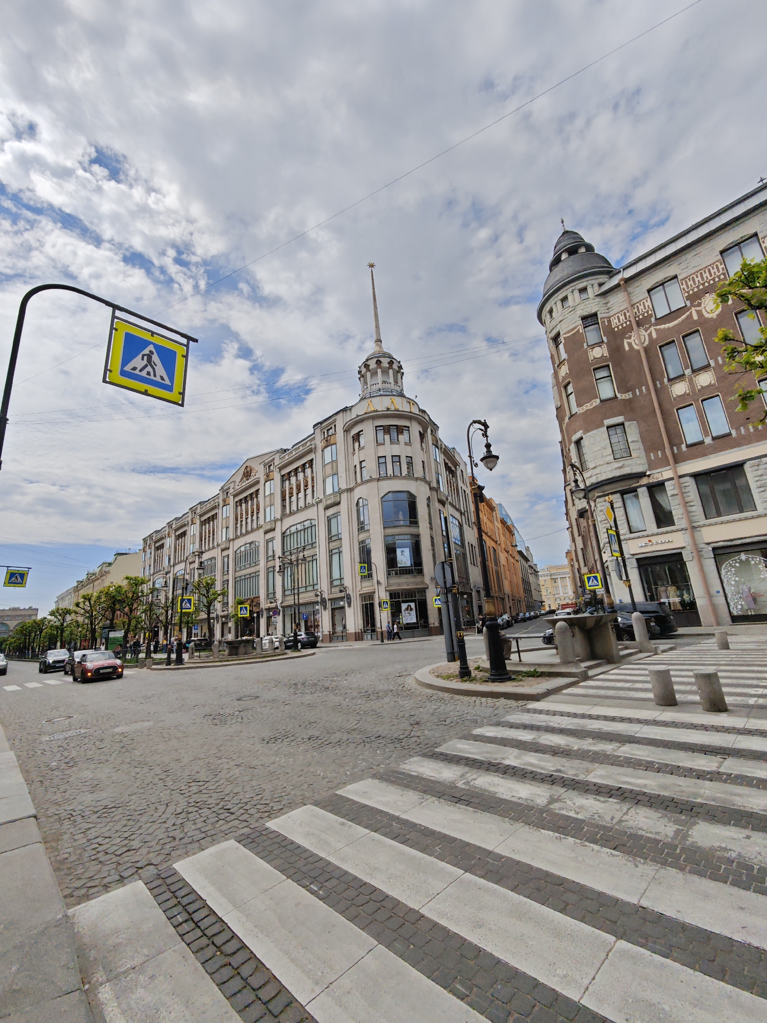 The image depicts a street scene in Saint Petersburg, Russia, with a focus on the intersection of Nevsky Prospect and Sadovaya Street. The foreground features a wide pedestrian crossing with white and dark grey stripes, paved with cobblestones and concrete slabs. Several concrete bollards are positioned to the right of the crossing.
On the left side of the intersection, a street sign indicating a pedestrian crossing is visible, mounted on a black pole. Several cars, including a red sedan and a black SUV, are moving through the intersection. The street itself is paved with cobblestones.
Dominating the background are two large buildings. The building on the left is a light-colored, multi-story structure with a distinctive spire topped with a star. The building on the right is a darker, brown building with ornate architectural details and multiple windows. Trees line the sidewalk between the buildings. The sky is cloudy with patches of blue. The scene appears to be a sunny day with moderate cloud cover. There are no discernible individuals engaged in any specific actions beyond driving and walking in the background.