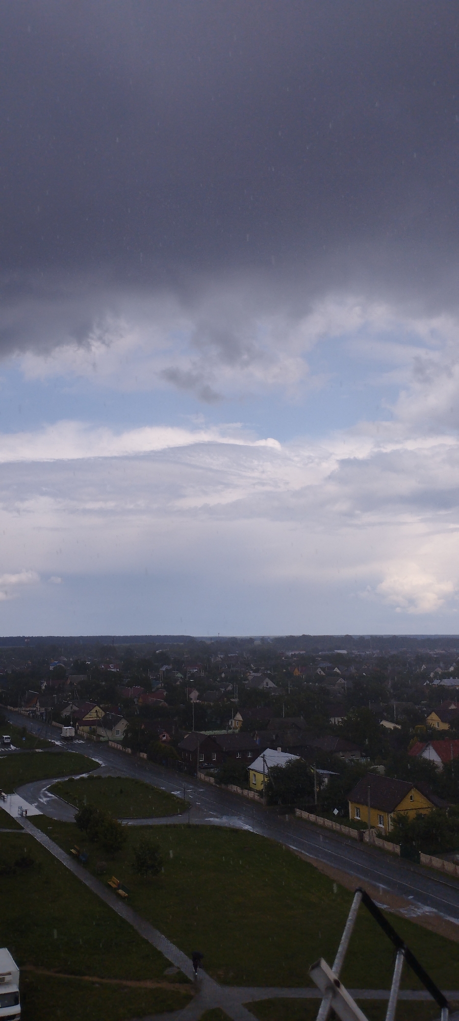 An aerial, vertical, outdoor, eye-level shot captures a broad view of Skidziel, Belarus, under a brooding, cloudy sky. The upper portion of the image is dominated by dark, heavy clouds, with lighter, more scattered clouds beneath them. The sky transitions to a pale blue near the horizon, where it meets a dense, dark line of trees that defines the edge of the town.
Below the tree line, the landscape unfolds into a sprawling residential area with numerous houses, primarily single-story structures with pitched roofs in various colors. Some houses have visible windows and chimneys. A paved road, wet from rain, winds through the settlement. A grassy area with benches and a walkway is visible in the foreground. A person is seen walking on the wet pavement, holding an umbrella. The scene appears to be experiencing rain, indicated by the wet surfaces and the visible streaks of precipitation in the air and on the ground. A white van is parked on the left side of the frame, and part of a white truck is visible at the bottom left. The overall impression is one of a damp, overcast day in a quiet, rural town.