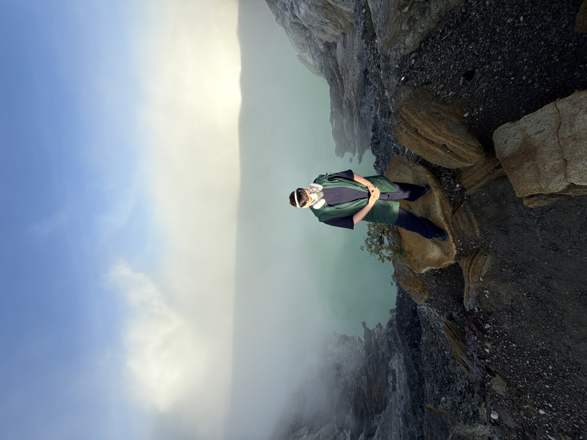 The image shows a person standing on a rocky edge near a body of water and a cloudy landscape, rotated 90 degrees. The person is wearing dark pants, a dark shirt, and a green jacket, with a respirator mask around their neck. The person's hands are clasped in front of them. The edge they are standing on consists of layered rocks and loose dark gravel. The water is a murky green color, and heavy fog or gas obscures the far side of the water. The sky is visible at the left of the image, transitioning from blue to white. The location is the Kawah Ijen volcano in East Java, Indonesia.