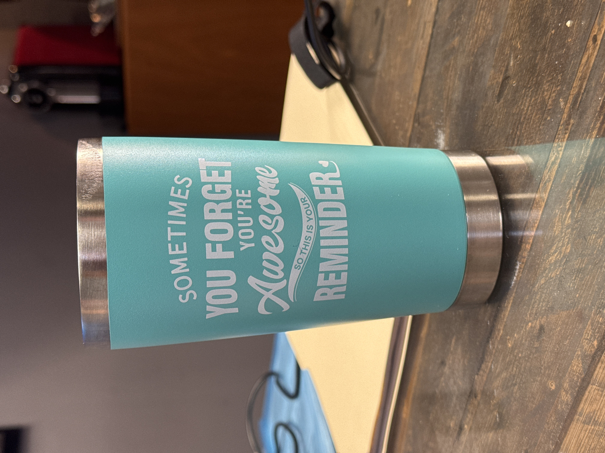 The image displays a teal-colored insulated tumbler resting horizontally on a dark, textured wooden surface. The tumbler features brushed metallic rims at both its top and bottom. Prominently printed on its side in white lettering is a motivational message: "SOMETIMES YOU FORGET YOU'RE Awesome SO THIS IS YOUR REMINDER". The word "Awesome" is rendered in a stylish, flowing script font, while the phrase "SO THIS IS YOUR REMINDER" is slightly smaller and also in a script font, accompanied by a decorative swoosh.
The wooden surface, likely a table or countertop, exhibits distinct grain patterns and appears somewhat worn or dusty in places. In the blurry background, various out-of-focus elements can be discerned, including a reddish object, a dark item with metallic details, and a light-colored wall or furniture, all indicative of an indoor setting. There is no specific activity occurring in the photograph; the tumbler is static. The overall scene suggests a casual indoor environment. The provided location context for this image is Apetatitlán, Mexico.