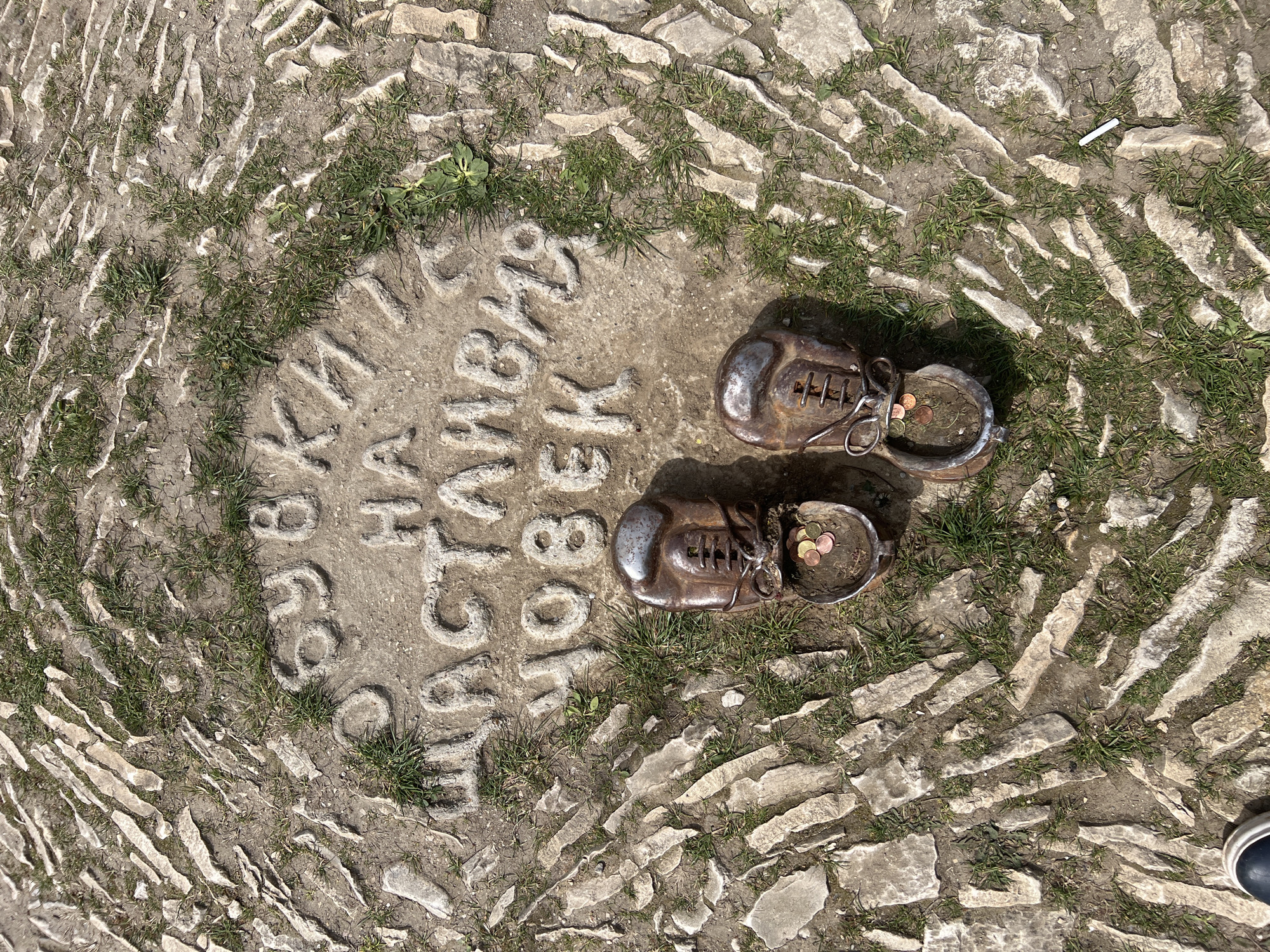The image depicts the "Shoes of Dancho Yordanov", a memorial in Lukovit, Bulgaria, set into a cobblestone surface. A circular inscription in Bulgarian surrounds two bronze shoes, each containing coins. A cigarette butt is visible near the inscription. The cobblestones are irregularly shaped and interspersed with grass and dirt. A small portion of a shoe is visible in the bottom right of the image.
