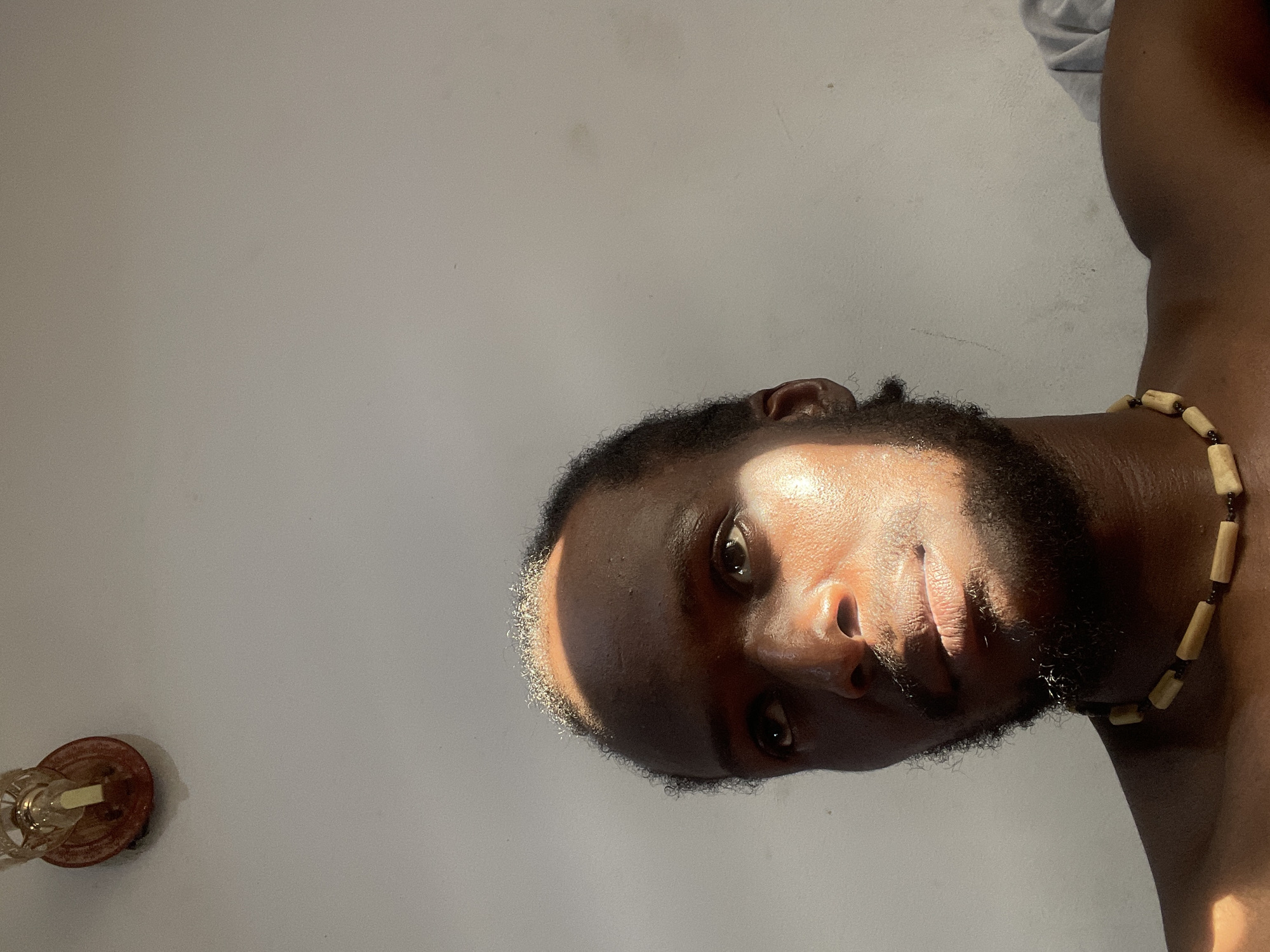 The image shows a close-up, rotated view of a Black man's face and upper body, against a plain, light-colored wall. He is wearing a necklace made of light-colored beads. The lighting suggests it is daytime, with a strong light source casting shadows across his face, highlighting his features and creating a dramatic effect. The sunlight appears to be coming from the side and slightly from above. The man has dark hair and a beard. His expression is neutral, and his eyes are looking directly at the camera. In the lower left corner, a part of a decorative lamp or light fixture is visible. The setting appears to be indoors, given the wall and the indoor fixture. No text is visible in the image. The location context is Abuja, Nigeria.