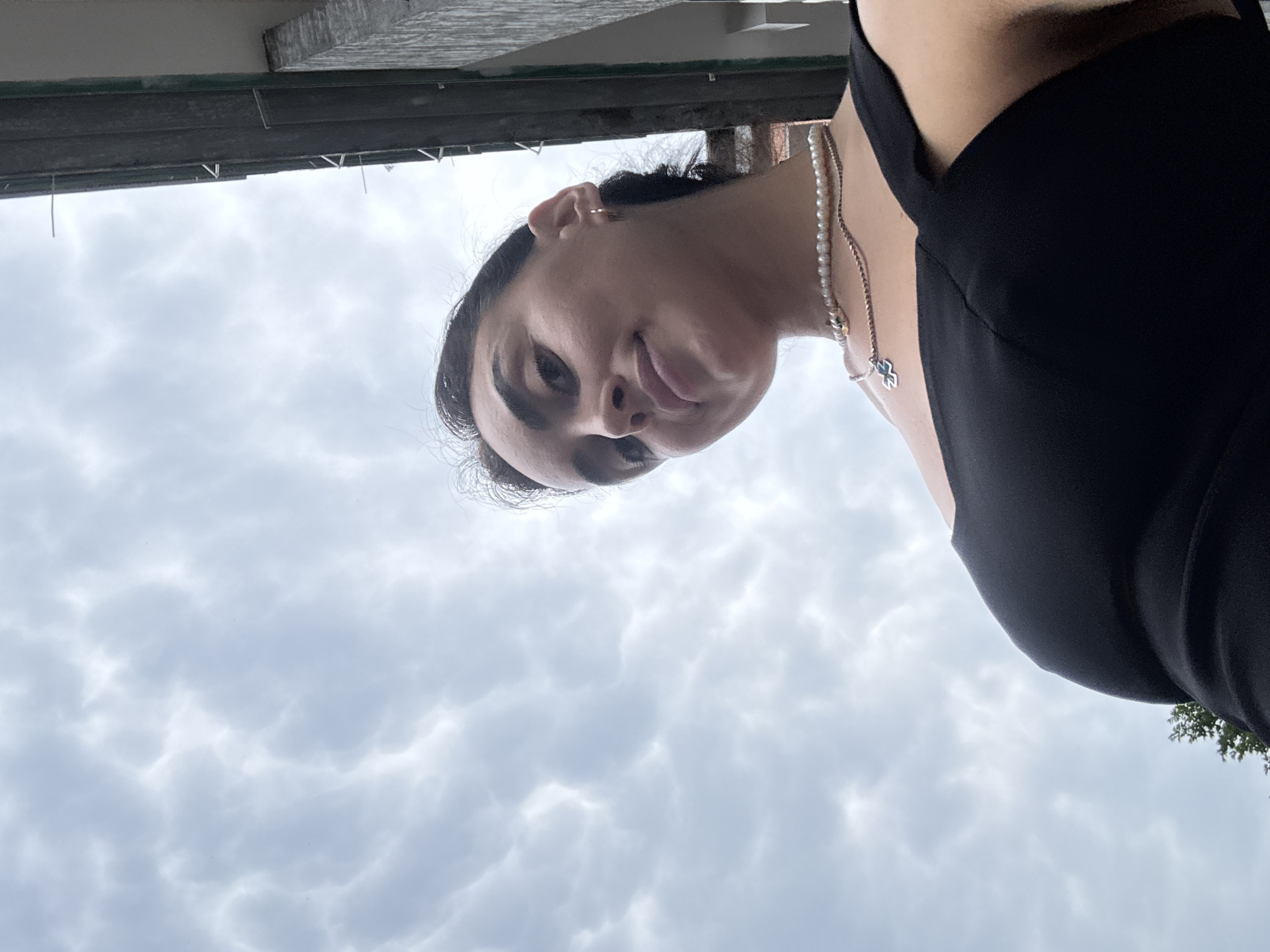 A low-angle shot shows a young woman with dark hair and a black dress, wearing a pearl necklace and a pendant. She is looking upwards, with a building and a cloudy sky in the background. The building appears to be made of concrete and metal, with some kind of railing or trim along the top. The clouds are dense and gray, suggesting an overcast day.