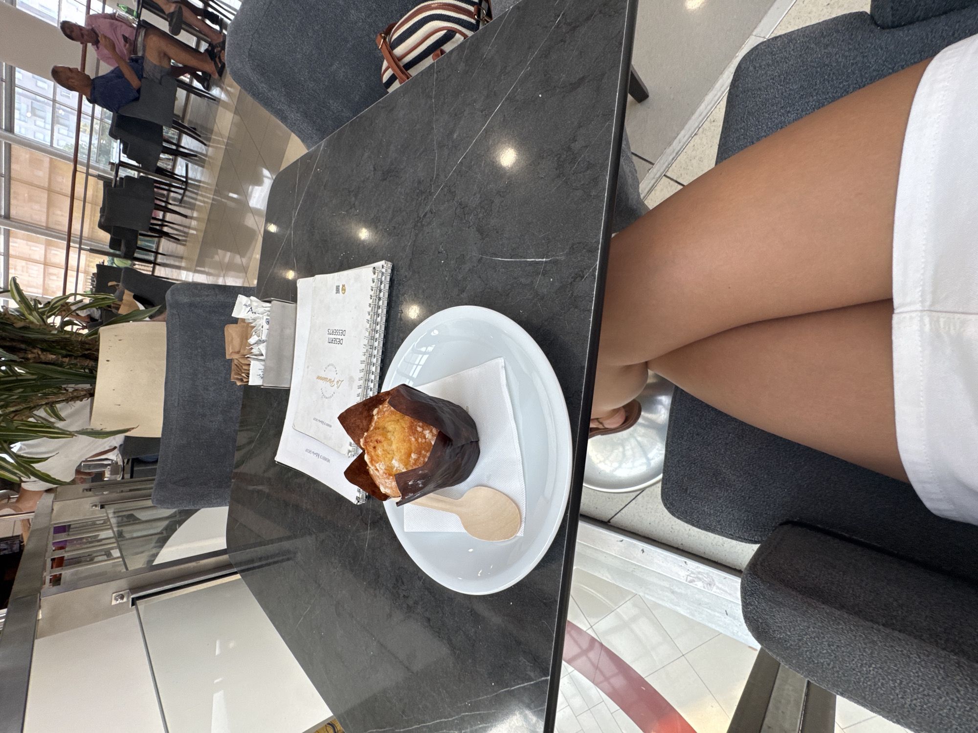 This is a high-angle, close-up shot of a person's legs crossed at the knee, sitting at a dark marble table. The person is wearing white shorts. On the table, there is a white plate with a muffin in a brown wrapper and a small wooden spoon. A spiral-bound notebook with text is also visible. In the background, there are other patrons sitting at tables and chairs in what appears to be an indoor cafe or restaurant. Through a large window, an outdoor scene with buildings and plants can be seen. The lighting suggests it is daytime. The image is oriented vertically.